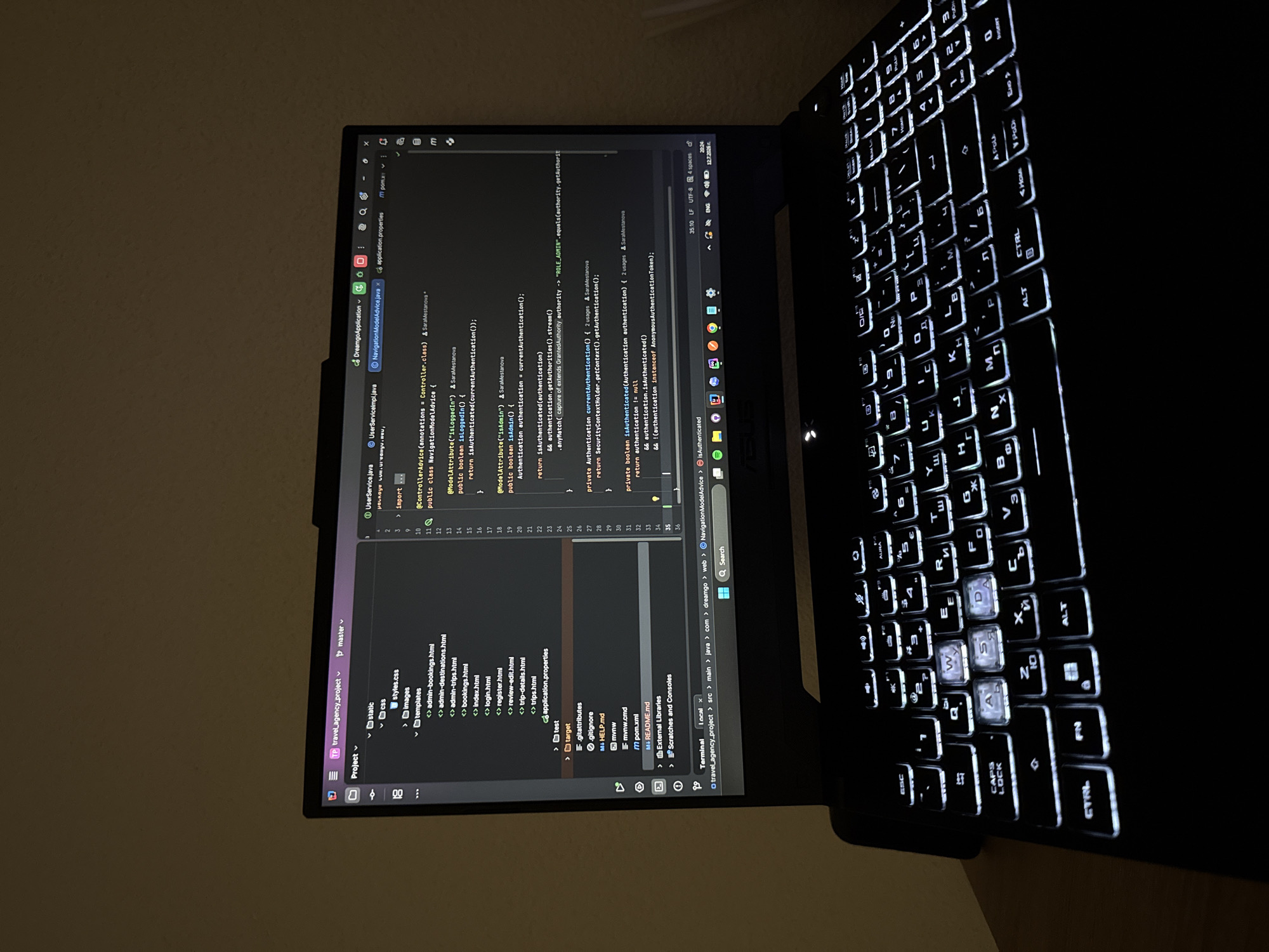 A laptop is positioned on a wooden surface, likely a desk or table, in a dimly lit room. The laptop screen displays a code editor with open files and project navigation. The keyboard is illuminated with white backlighting, highlighting keys such as WASD, Ctrl, Alt, and the function keys. The overall scene suggests a work or study environment. The image does not contain information about the city of Krushev Dol, Bulgaria.