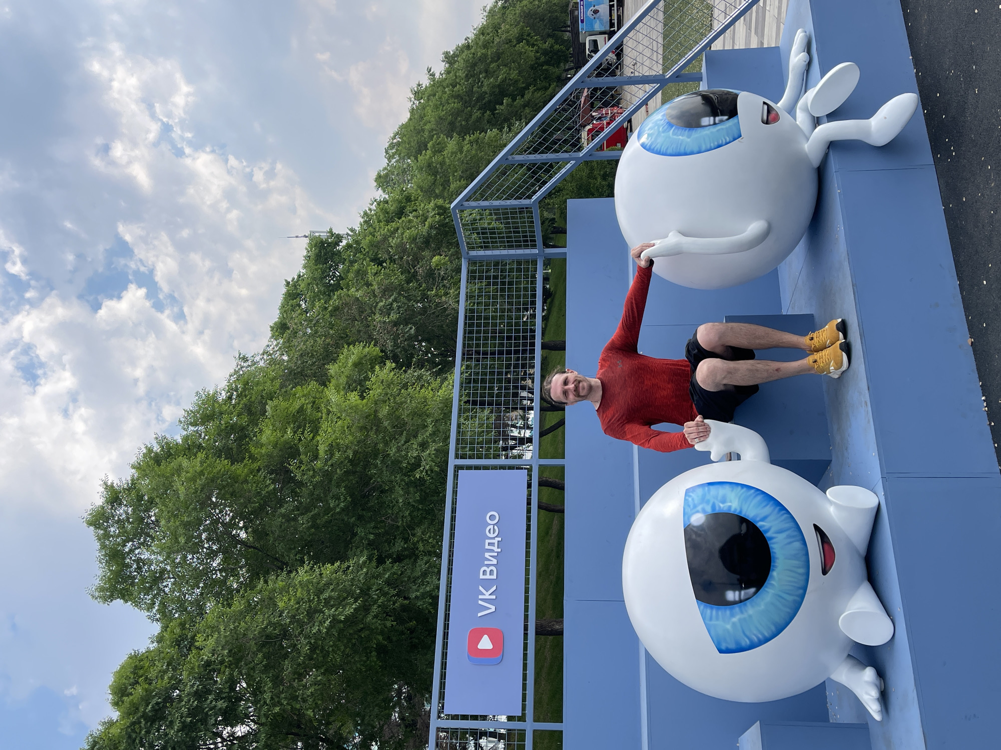 A man in a red shirt and black shorts sits between two large, cartoonish eye sculptures with bright blue irises. The sculptures are white with cartoonish smiles. The man is leaning on the left sculpture with his left hand, and his right hand is resting on his knee. The left sculpture is sitting down with its legs extended forward, and its left hand is raised as if waving. The right sculpture is also sitting down with its legs extended forward and to the side, and its right arm is bent at the elbow. Behind them is a blue structure with a sign that reads "VK Видео" with a play button icon. The background consists of lush green trees and a cloudy sky. The scene appears to be an outdoor public space, possibly a park or an event. Based on the "VK Видео" sign, the location is likely related to the VKontakte social media platform.