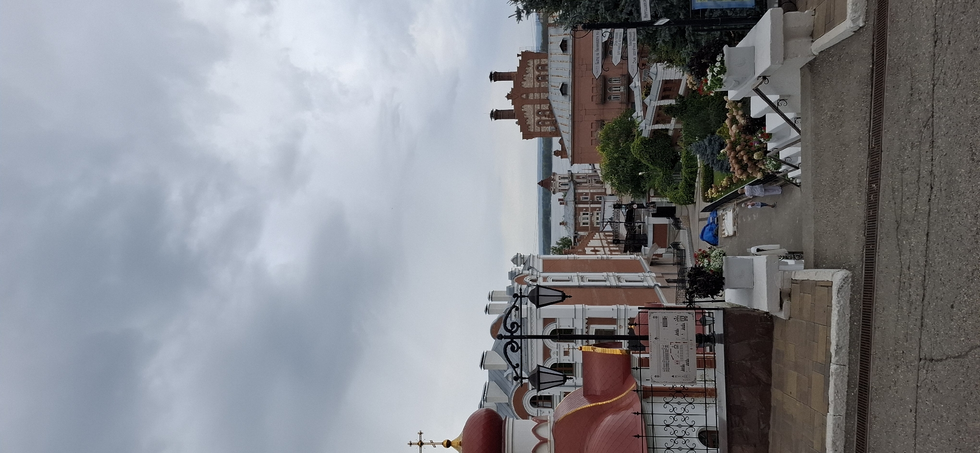 The image shows a view of a city in Samara, Russia, on a cloudy day. In the foreground, a steep concrete staircase descends from the viewer's perspective, with a metal grate running along its length. To the left, a white wall and a tiled walkway are visible, leading to a red-domed building with a golden cross, likely a church. Ornate black lampposts stand along the walkway.
Further down the walkway, several multi-story buildings with red brick and white trim line a street. Some of these buildings have distinctive architectural features like arched windows and decorative chimneys. Green trees and flower beds are interspersed among the buildings, adding color to the scene.
In the mid-ground, two small figures, appearing to be a woman and a child, are walking down a paved pathway. A blue tarp is spread on the ground near them.
In the background, a wide body of water, likely a river or bay, is visible. Several more buildings, including a prominent red brick structure with multiple chimneys and a smaller building with a domed tower, are situated along the waterfront. Several street signs with Russian text are mounted on poles in the mid-ground. The sky is overcast with heavy gray clouds.