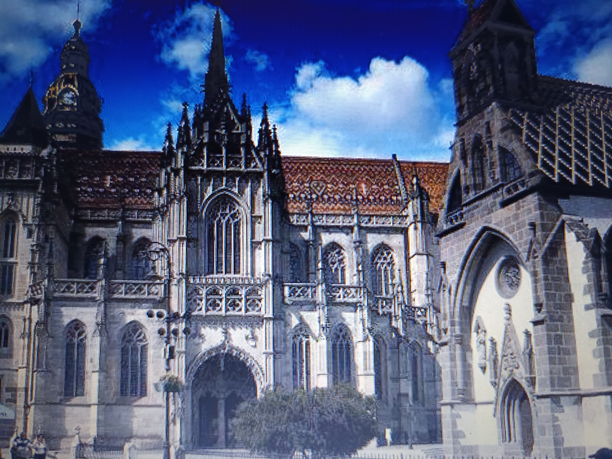 The image displays a low-angle, wide shot of St. Elizabeth's Cathedral in Košice, Slovakia, under a bright blue sky with scattered white clouds. The Gothic architecture of the cathedral dominates the frame, with its intricate stone carvings, pointed spires, and reddish-tiled roofs. The facade features large stained-glass windows, arched doorways, and decorative stonework. To the left, a clock tower with a dome is visible. In the foreground, a dense green tree partially obscures the entrance. A few indistinct figures of people can be seen near the bottom left of the image, and a low wall or fence runs along the bottom. The overall impression is a grand and historic religious building.
