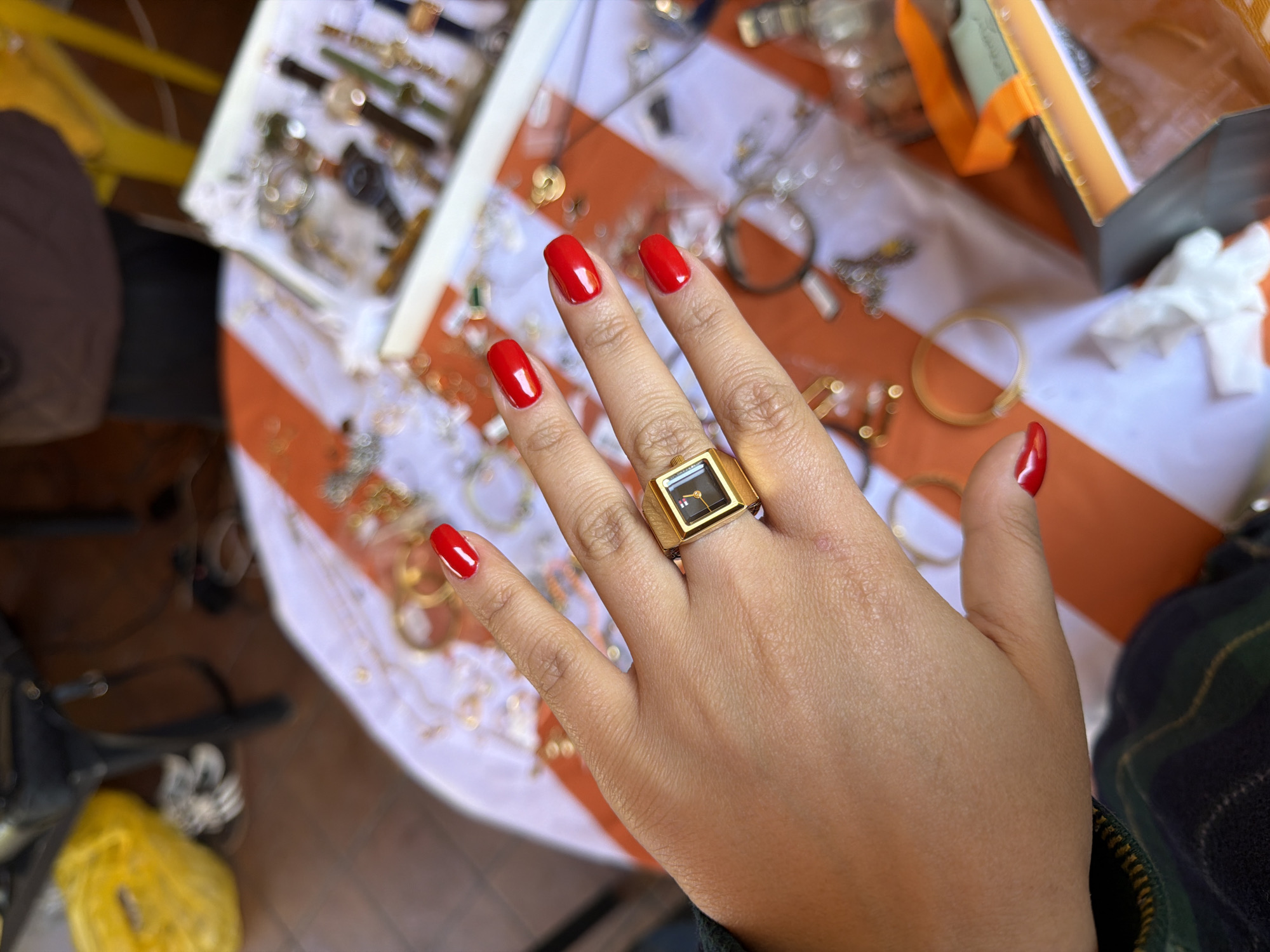 A right hand with red nail polish is the main subject. A large gold ring with a square black watch face is on the ring finger. The hand rests on a table covered with a white cloth with orange stripes. Displayed on the table are various pieces of jewelry, including watches and necklaces. A white jewelry tray and a partially visible orange box are also present. The overall setting suggests a jewelry market or shop. The location in Yerevan, Armenia is not evident from the image itself.