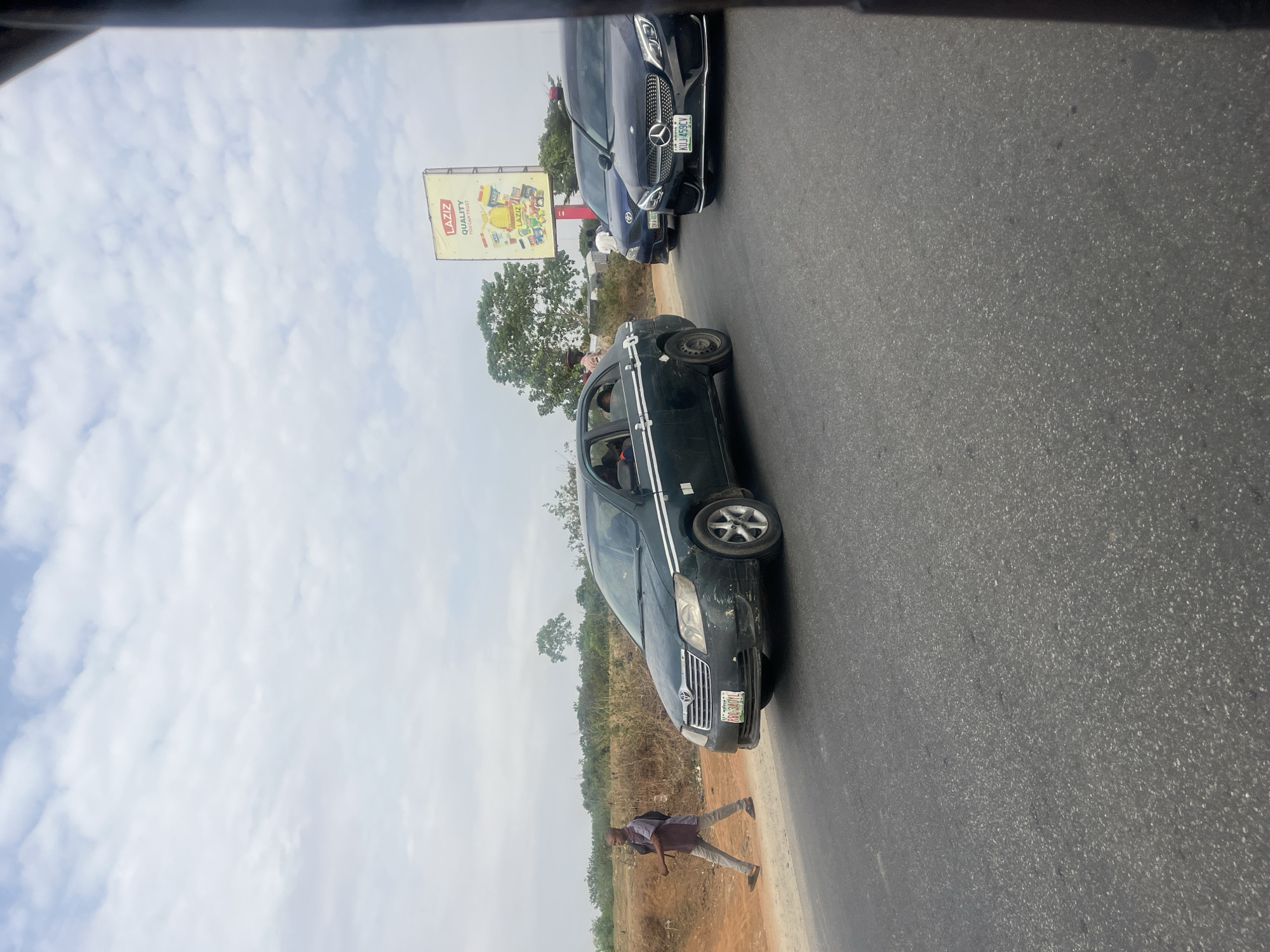 The image shows a section of road near Karmajiji, Nigeria. A dark-colored sedan with a spare tire mounted on the front left wheel is parked on the side of the road. A man in a brown shirt and light-colored pants walks along the shoulder of the road near the sedan. A dark-colored Mercedes-Benz with license plate KUU 459 CY is parked in the opposite lane. A billboard for LAZIZ is visible. The sky is overcast.