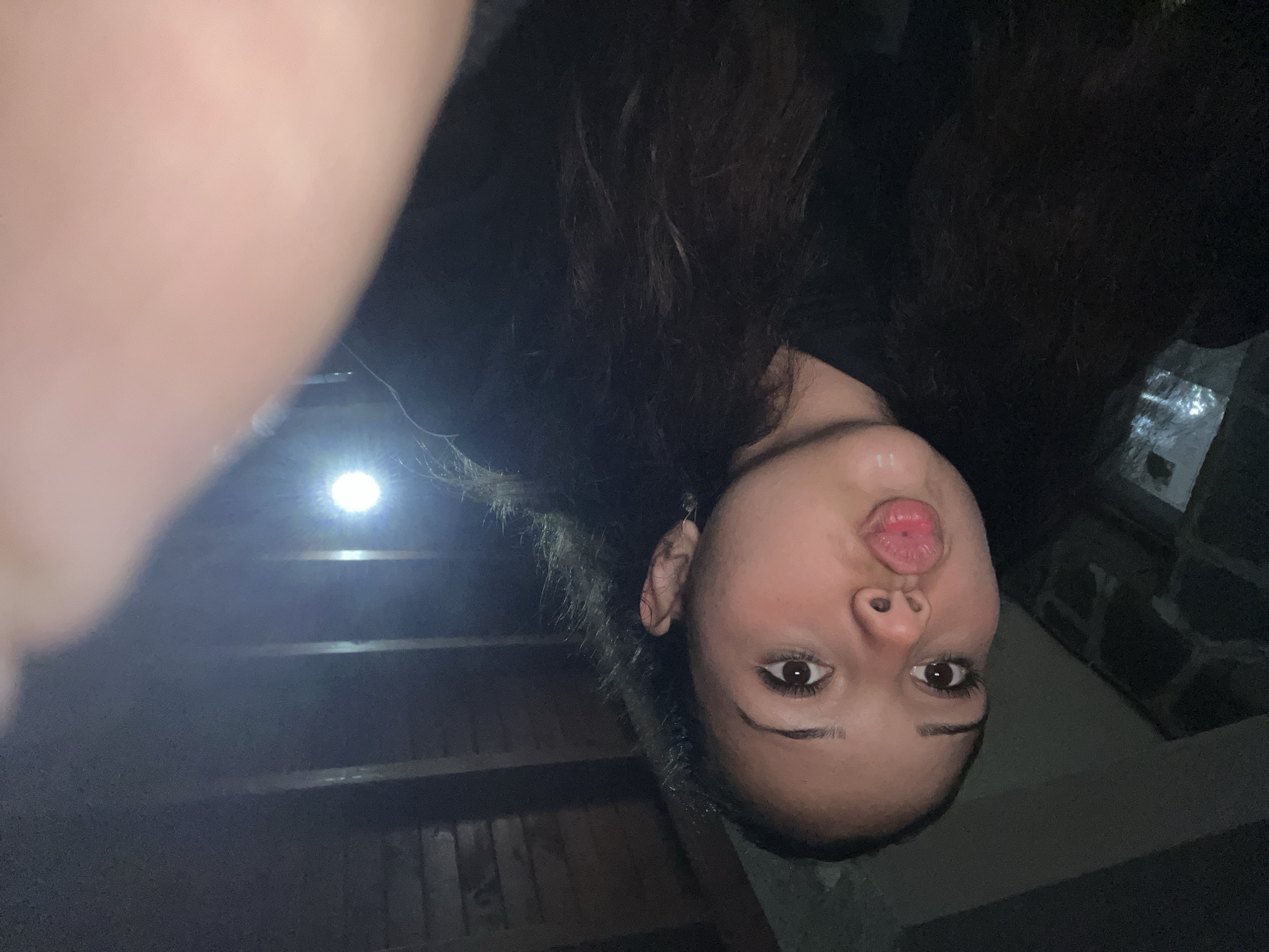 A young woman with dark hair is making a kissing face at the camera, which is held close to her. She is wearing a dark shirt. The background appears to be indoors with wooden beams and stone walls visible. A bright light source is present in the background. The image is grainy and underexposed, suggesting it was taken in low light conditions. The location is Zlatograd, Bulgaria.