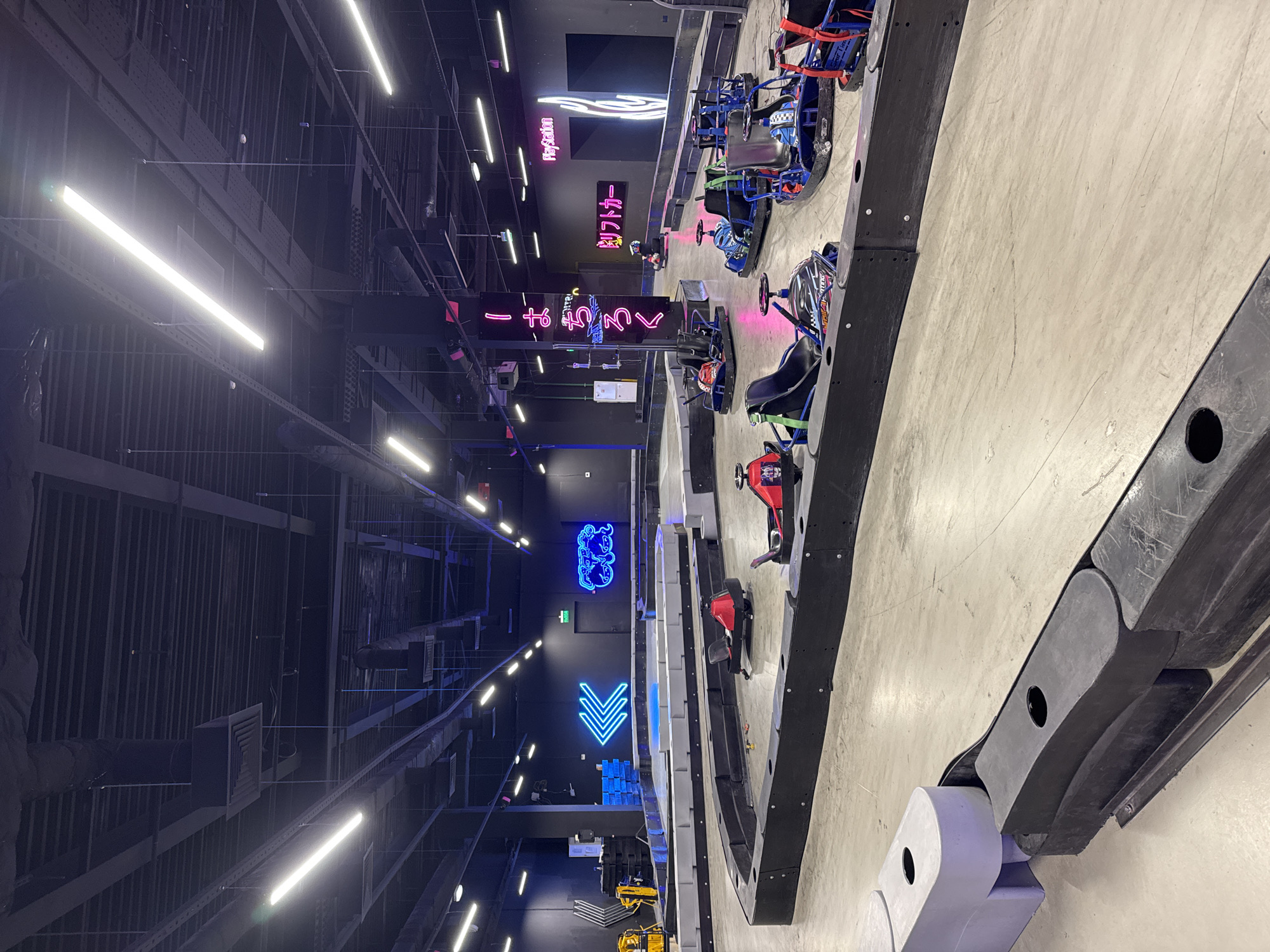 The image depicts an indoor go-kart track. The track is enclosed by black and gray barriers. Several go-karts are visible on the track, some appearing to be red and others blue. The environment is dimly lit with artificial lighting, including long fluorescent tubes and neon signs. One neon sign on the back wall features Japanese characters in pink and white. Another neon sign displays the PlayStation logo. The ceiling is dark and industrial, with visible ductwork and lighting fixtures. The floor is concrete. There are no people clearly visible in the foreground or midground, though a figure may be present in one of the go-karts in the distance. The scene appears to be a commercial entertainment venue. The location is identified as Sovkhoza imeni Lenina, Russia, suggesting this is an entertainment facility in that area.
