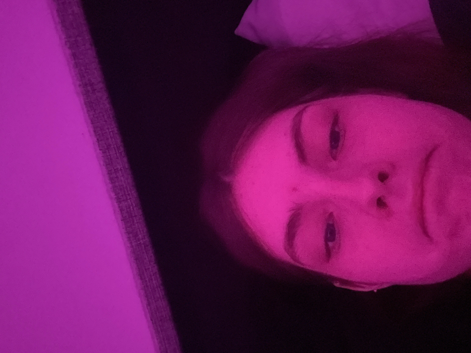 A close-up, rotated image shows a person's face illuminated by a purple light. The person has fair skin, dark hair, and visible eyes, nose, and lips. To the left, a textured gray surface is partially visible. The background is dark, suggesting a dimly lit room. No information suggests this image was taken in Saint Petersburg, Russia.