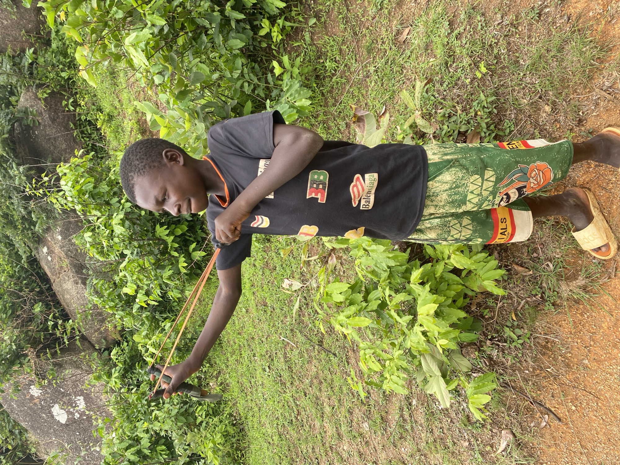 A young boy stands in Ganawuri, Nigeria, surrounded by lush greenery. He is actively drawing back a slingshot, aiming towards a target outside the frame. He is clad in a black t-shirt with visible logos and green patterned shorts. He is also wearing sandals. The environment consists of grass, scattered foliage, and rocks.