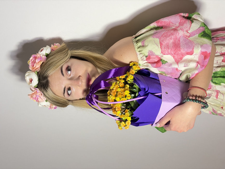 The scene is set in Budva, Montenegro, against a plain, light grey background. A young adult female with blonde hair, styled with a floral headband featuring white and pink artificial flowers, occupies the central frame. She wears an off-the-shoulder garment adorned with a large pink and green floral pattern. Her left hand holds a potted plant containing numerous small yellow flowers, presented in a decorative light and dark purple paper holder with a quilted texture and heart cutouts at the base, cinched with a purple ribbon forming a bow and straps. On her left wrist, she wears multiple bracelets, including beaded green and reddish-brown ones and a thin red braided cord. Her head is slightly tilted, and her gaze is directed towards the right side of the frame. Her lips are parted, showing dark pink lipstick. A distinct shadow is cast on the wall to the upper right of the subject.