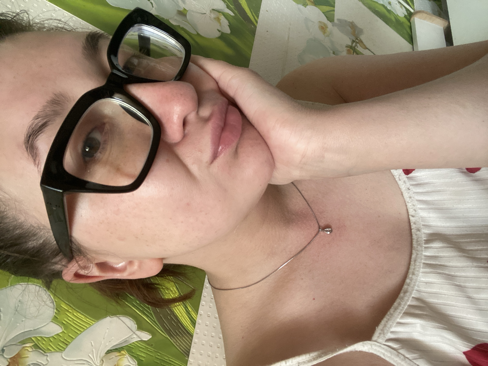 A close-up, slightly angled shot depicts a woman wearing large, black-framed glasses and a thin silver necklace with a small pendant. She rests her cheek on her hand, with her fingers near her mouth. The background is partially obscured but appears to be a surface with a floral design. The lighting is soft and diffused. There is no information to confirm the location in Chaikovsky, Russia.