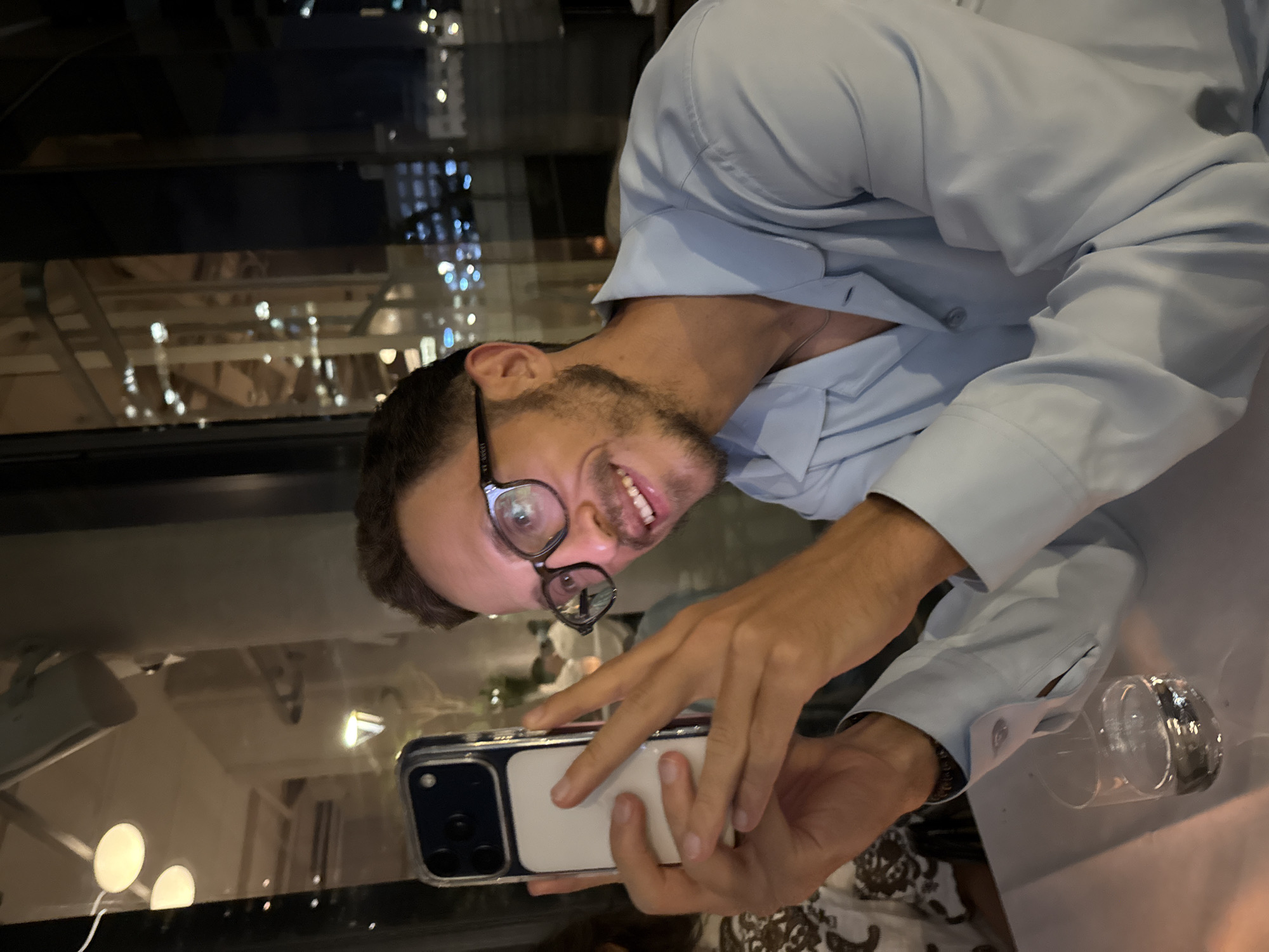 A young man wearing a light blue collared shirt and glasses is taking a selfie with his phone in a restaurant in Tel Aviv, Israel. The background shows a window overlooking a city at night with blurred lights. He is smiling and holding his phone with both hands. There is a glass of water on the table in front of him.