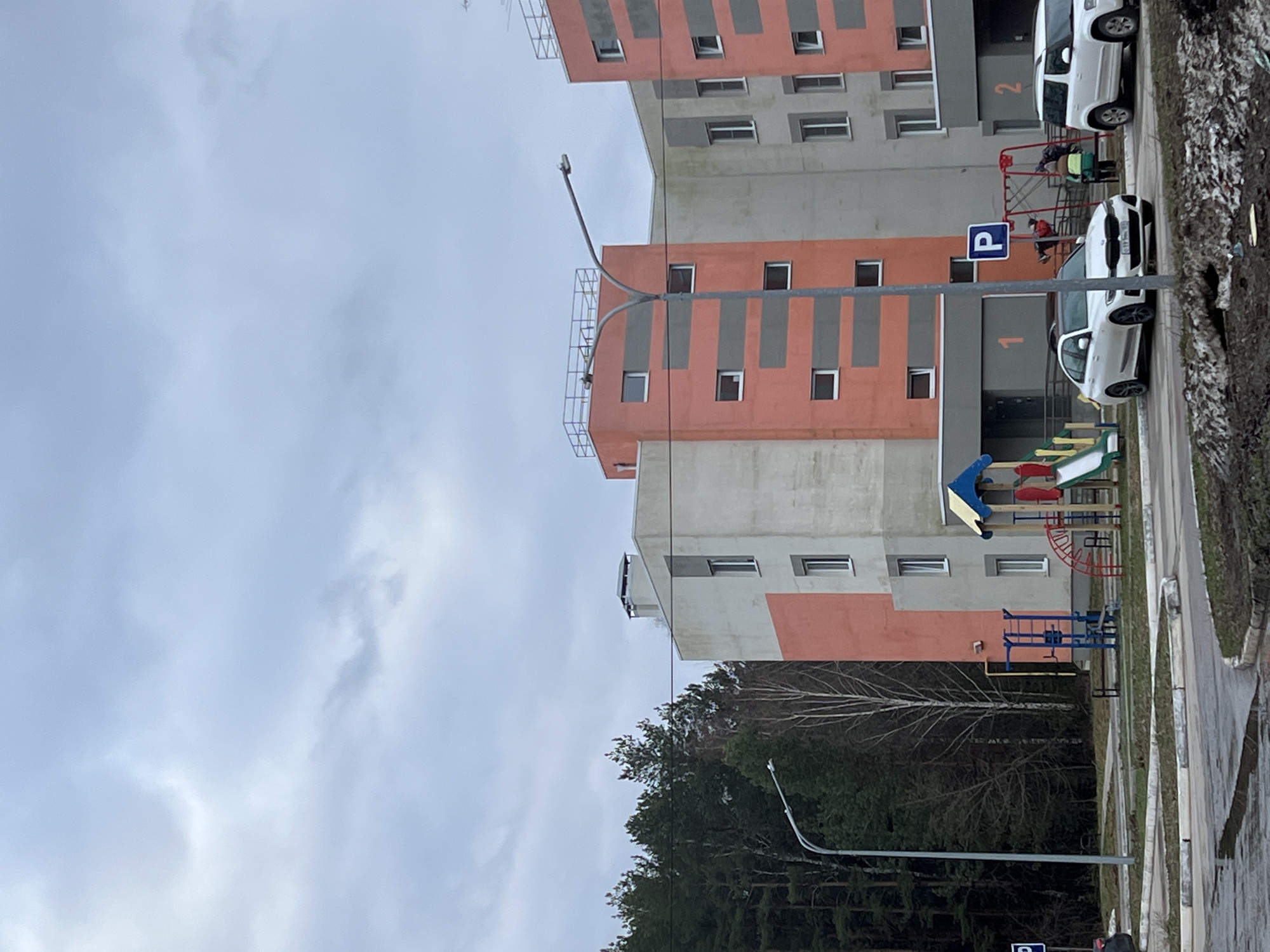 This daytime image captures a residential area in Chaikovsky, Russia, under an overcast sky. The scene features two modern multi-story residential buildings with facades painted in light grey/beige and salmon-orange, accented by darker grey vertical strips. Large numbers "1" and "2" are prominently displayed near the ground-level entrances of the respective buildings.
In the foreground, a colorful children's playground with blue, yellow, and red slides and climbing frames is visible. Adjacent to the playground, a paved road or parking area hosts several parked white vehicles, including a white SUV and a white BMW sedan. A blue square sign with a white "P" clearly marks a parking zone. A few people, including one person in a bright green/yellow jacket and another in red, are on or near the playground equipment.
A line of trees, including both bare deciduous trees and evergreens, borders the residential complex, hinting at a natural or wooded area nearby. Several streetlights are also present, with one in the foreground to the left and another further back. The sky is consistently cloudy and light grey, suggesting a dull, overcast day.