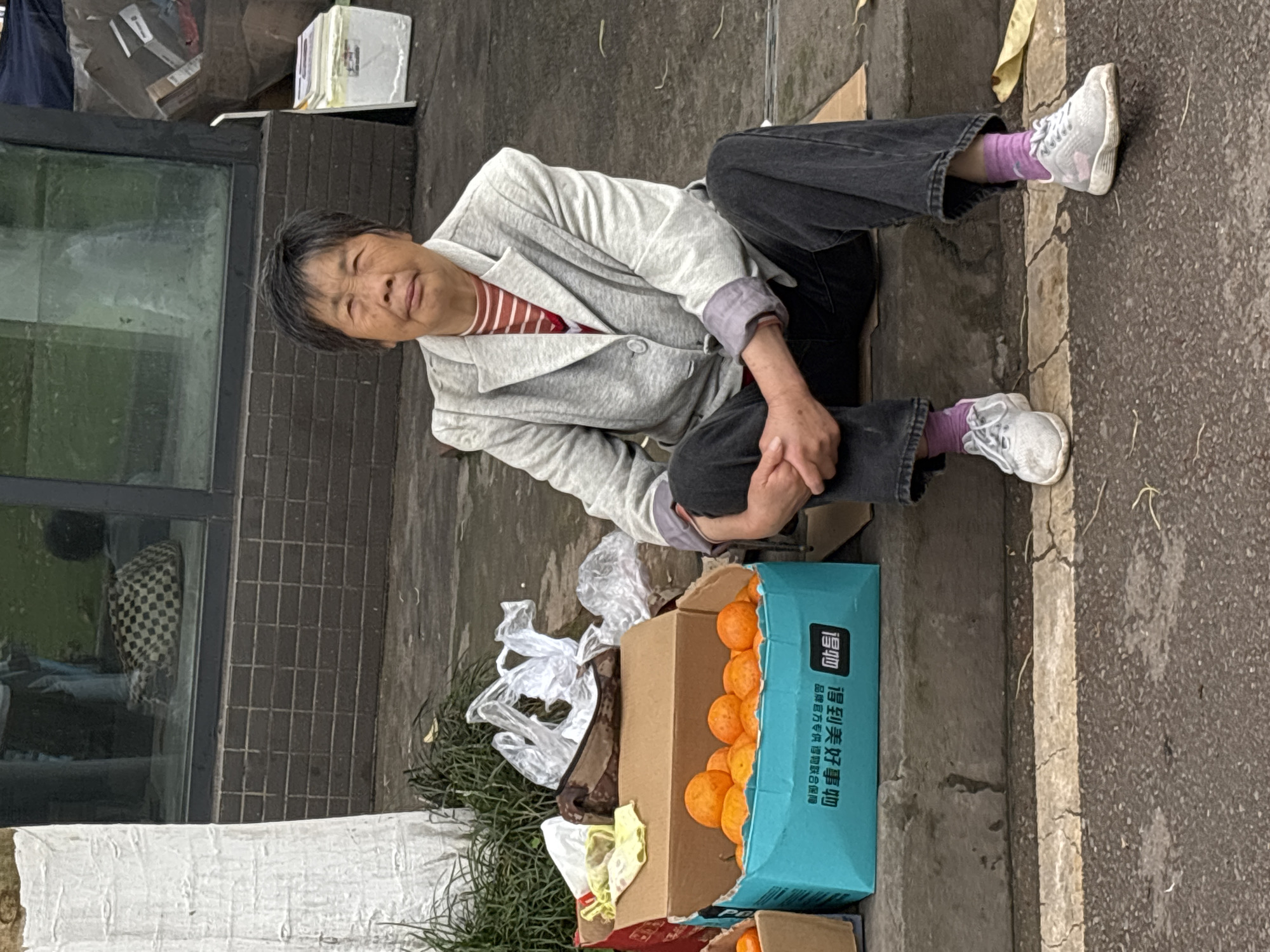 A middle-aged Asian woman sits on the ground near a building, with her legs extended and crossed at the ankles. She is wearing a light gray jacket, a red-striped shirt, dark cropped pants, pink socks, and white sneakers. A box of oranges sits next to her, along with some trash and vegetation. The background includes a gray wall, a window, and a street. The location is likely Jiangjin, China.