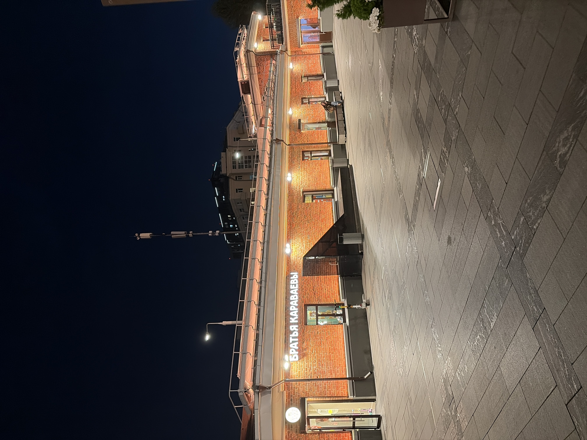 This is a nighttime, eye-level, medium shot of the exterior of the "Brat'ya Karavaevy" cafe in Moscow, Russia. The facade of the cafe is made of red brick and is illuminated by warm orange light from above and spotlights on the brickwork. A large neon sign above the windows reads "БРАТЬЯ КАРАВАЕВЫ". The cafe has large windows and two glass doors, one of which is open, revealing a brightly lit interior. A person is seated on a bench outside the cafe. The ground in front of the cafe is paved with large, gray stone tiles arranged in a patterned walkway. In the background, a dark night sky is visible with a tall cell tower and distant buildings with lights.
