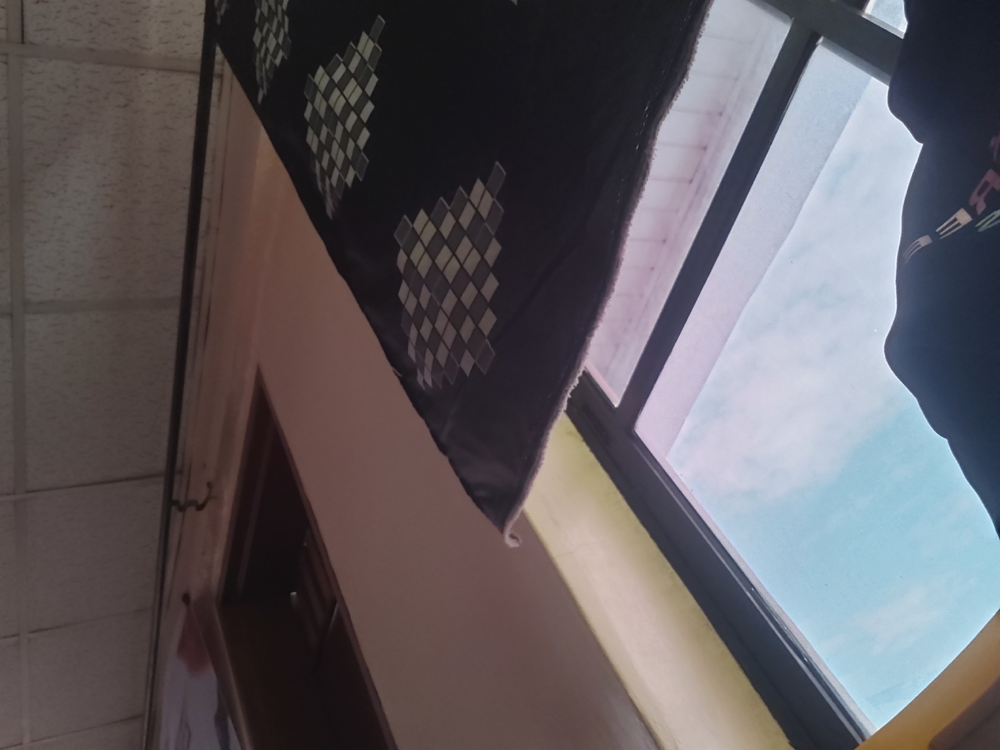 The image shows an interior view looking upwards and outwards. A black textile with a diamond pattern is draped over the top portion of a window frame. The window itself reveals a bright, light blue sky with faint clouds. Below the window, a dark silhouetted figure is visible, with a portion of a black garment with a graphic design showing. The walls appear to be a light beige color, and the ceiling has a textured, tiled appearance. The scene does not provide any specific visual cues to determine its location within Jos, Nigeria.