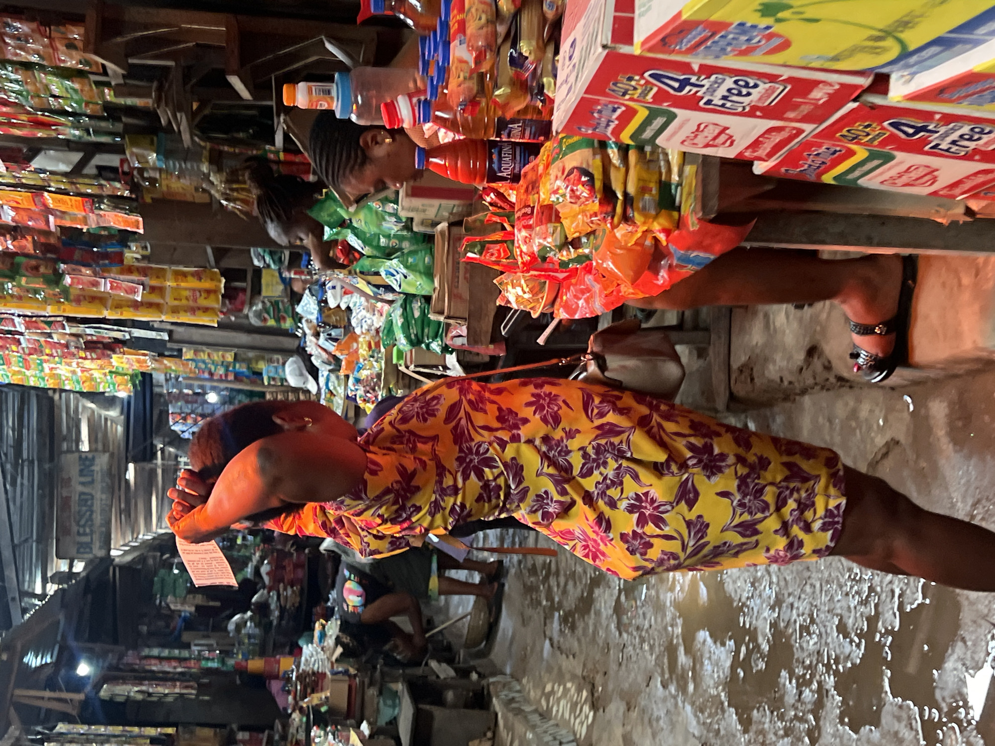 A busy marketplace scene in Nkpor, Nigeria, is depicted with a woman in a yellow and purple floral dress holding a piece of paper. She is positioned in the foreground, facing away from the camera, with her right hand raised near her head as she examines the paper. The market stalls are laden with various goods, including packets of food items, bottles of drinks, and other products stacked on shelves and tables. The ground appears wet and muddy, suggesting recent rain or a water source within the market. Other individuals are visible in the background, engaged in activities typical of a marketplace, such as tending to stalls or shopping. The overall atmosphere is one of commercial activity and daily life within the Nigerian city.