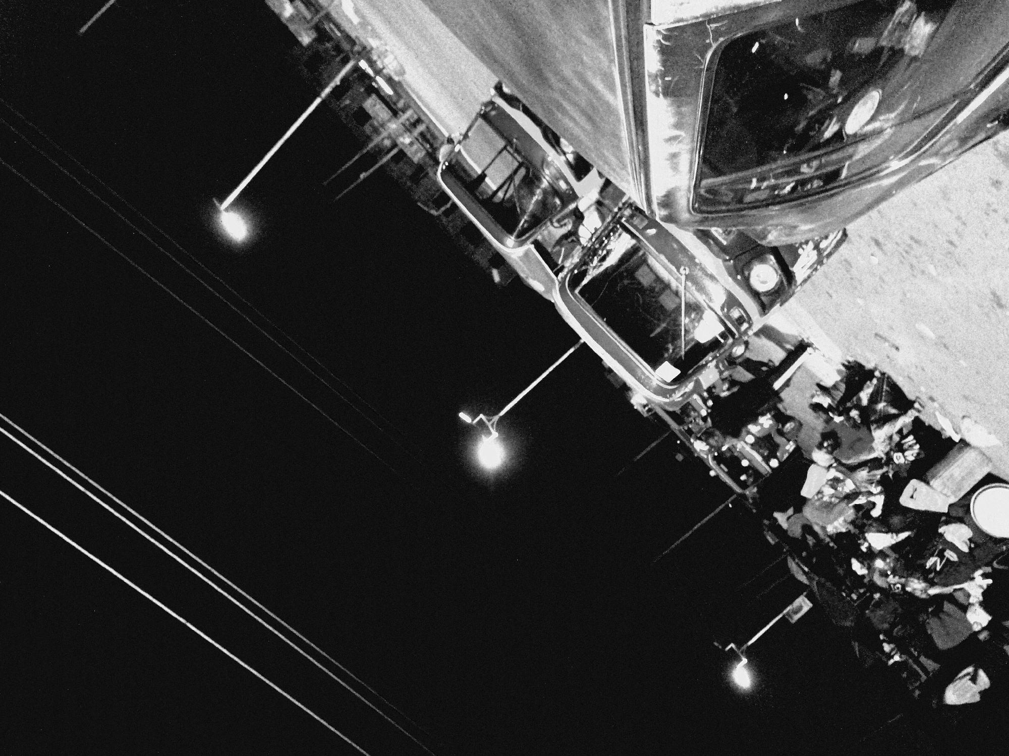 This is a black and white, high-angle, night-time photograph of a street scene in Bukuru, Nigeria, with an apparent pile of goods or luggage on the left. Power lines are visible overhead, stretching diagonally across the frame. A street light illuminates the scene from the upper right. Several auto rickshaws, commonly known as 'keke' in Nigeria, are parked or moving. In the foreground on the right, a dark-colored vehicle, possibly a van or SUV, is parked, with its side panel and window reflecting the ambient light. The ground appears to be a paved road or street. The overall impression is one of urban activity at night, with modes of transport and goods suggesting a commercial or transport hub.