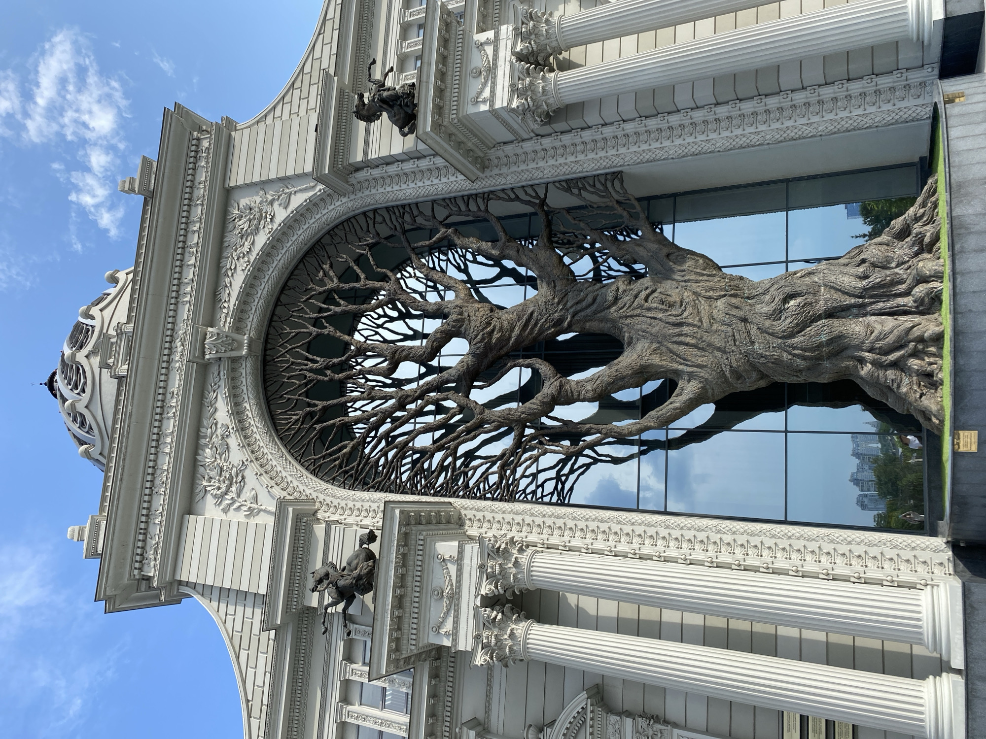 The image depicts the National Museum of the Republic of Tatarstan, located in Kazan, Russia. The museum is characterized by its grand neoclassical architecture, featuring columns, ornate carvings, and a dome. Prominently displayed in front of the building is a massive, sculptural representation of a bare tree with intricate branches that fill a large arched window. The tree sculpture appears to be made of a textured, dark material, possibly metal or resin, and its roots spread out at the base. The building's facade is light-colored, contrasting with the dark tree sculpture. Two equestrian statues are visible on pedestals on either side of the arched entrance, adding to the building's majestic appearance. The sky above is blue with scattered clouds. Reflections of the sky and nearby buildings are visible in the large glass windows behind the tree sculpture. The overall scene is static, with no people or animals actively engaged in any discernible actions.