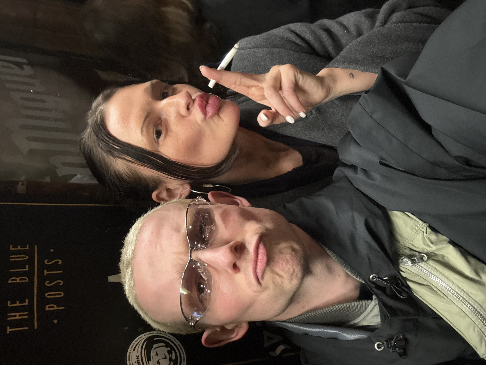 A selfie photograph shows a man and a woman indoors. The man has a light-colored crew cut, wears glasses, and a layered jacket. The woman has dark hair and is holding a lit cigarette while making a kissing face. She wears a gray top, and a small tattoo is visible on her left wrist. A sign for "THE BLUE POSTS" is partially visible in the bottom left corner, suggesting the location is The Blue Posts pub in Soho, London, United Kingdom.