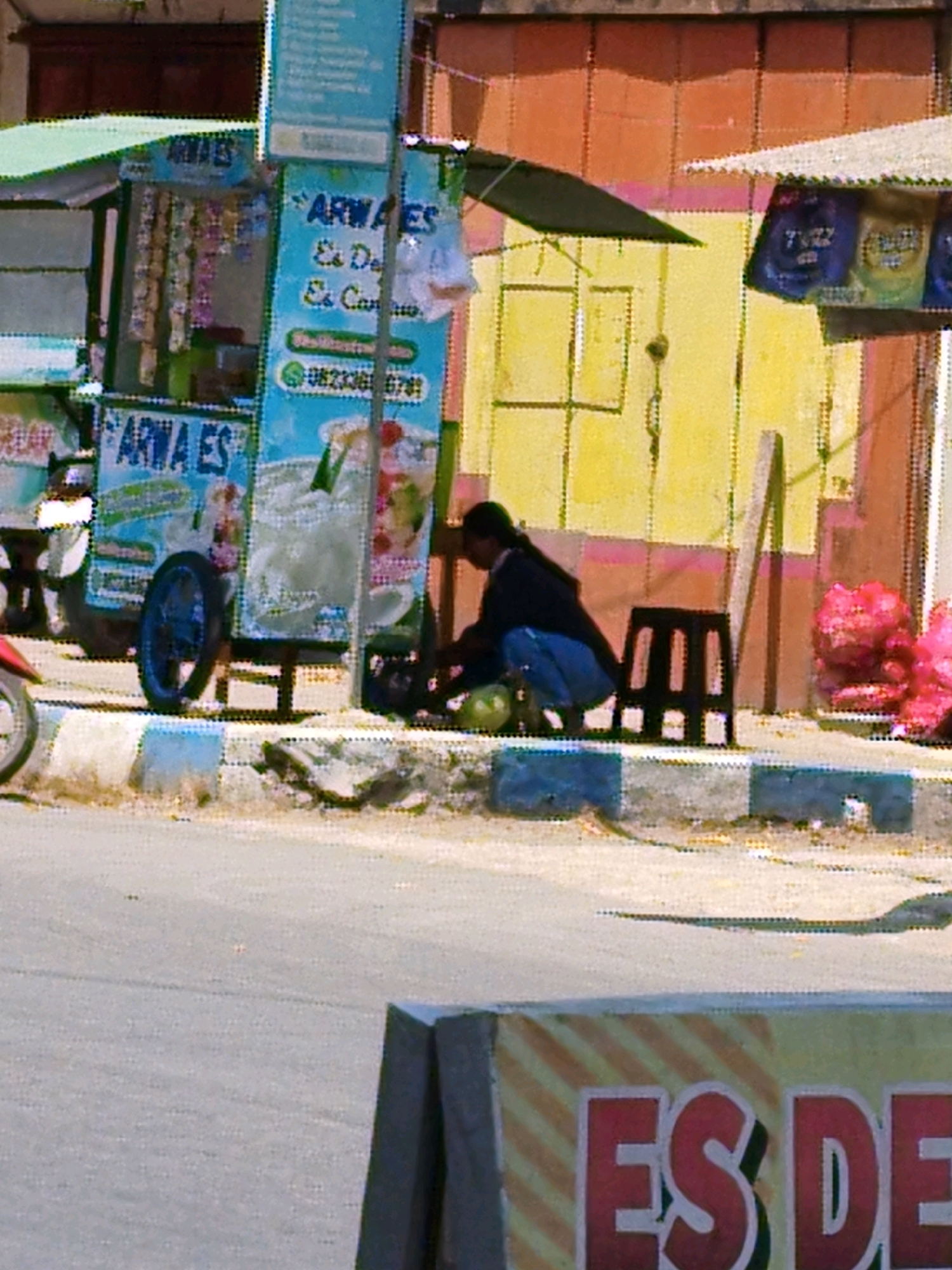 In Arjowilangun, Indonesia, a street scene unfolds with a focus on a mobile food cart selling "Arwa Es". The cart displays a colorful sign with the brand name and images of various drinks. Beside the cart, a person, likely the vendor, is crouched down, tending to the offerings. In the background, a yellow wall with orange accents forms the backdrop, with a glimpse of a store awning and shelves stocked with bagged items. A black stool sits near the vendor, and a pile of pink balloons is visible to the right. The scene is situated on a street with a sidewalk bordered by a curb. The overall impression is that of a typical street vendor operation in a local Indonesian setting.