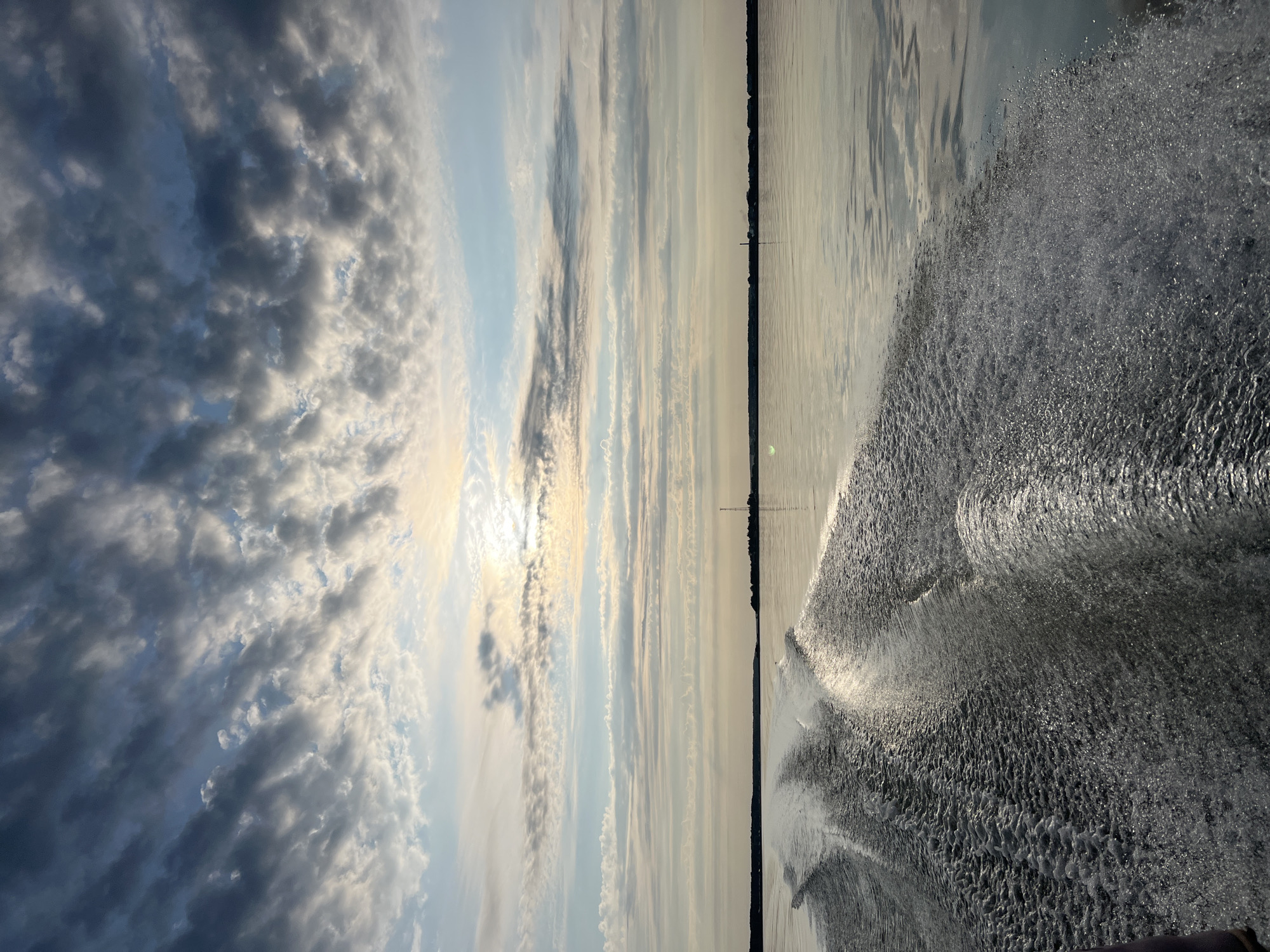 The image depicts a dynamic seascape from the perspective of a moving boat. In the foreground, prominent white wakes and turbulent water are visible, indicating rapid movement. The water surface is largely calm beyond the wake, reflecting the sky. The horizon line is low, featuring a flat, distant shoreline dotted with trees and structures, including a tall antenna. The sky is filled with a dramatic display of clouds, with streaks of white and grey against a pale blue and yellow sky, suggesting either sunrise or sunset. The sun itself is partially obscured by clouds, casting a diffused light. The scene is serene and evokes a sense of travel and nature. The location is identified as Novopichugovo, Russia.