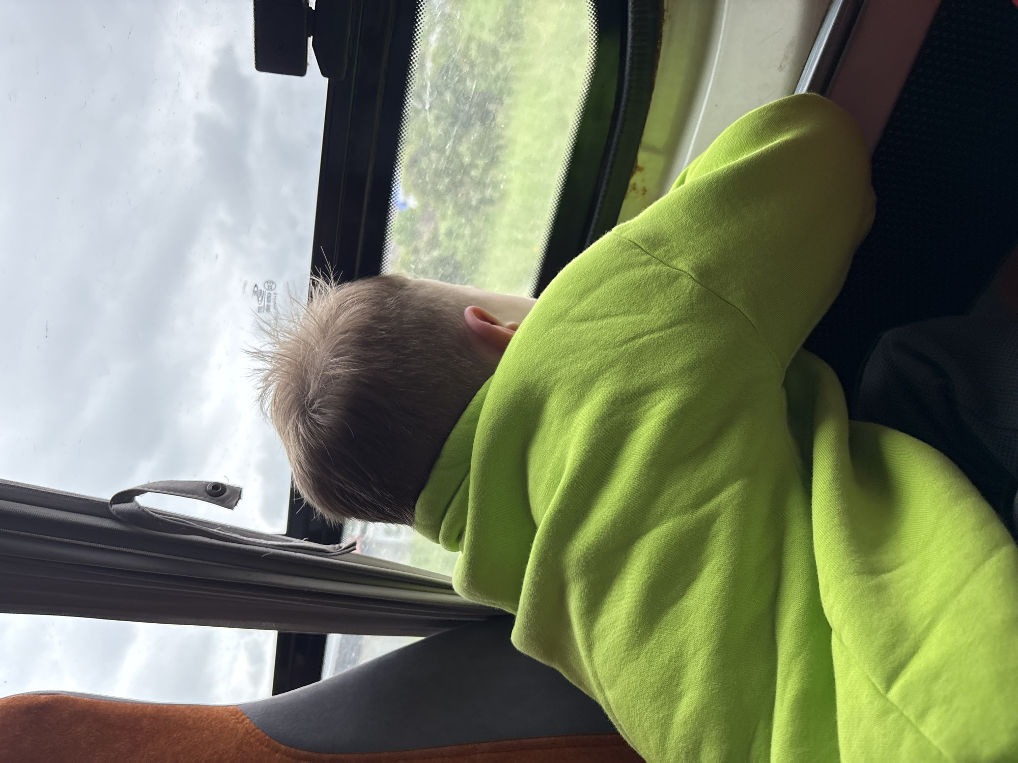 The image captures a scene from within a vehicle located in the city of Chaikovsky, Russia. A child, viewed from the posterior aspect, occupies the foreground. The child has short, light-colored hair and wears a vibrant lime green hooded sweatshirt, characterized by visible fabric folds. Leaning forward, the child rests their forearms on the vehicle's window sill or lower frame, intently observing the exterior landscape through a dark-framed window. The window glass exhibits subtle smudges. To the left of the child, a section of an orange seat with dark grey trim is visible. A grey fabric curtain, fastened back by a loop with a dark snap, hangs vertically alongside the left window frame. The exterior view consists of an overcast sky, presenting a uniform light grey tone, above a broad, green, presumably grassy, field. A small, dark device is affixed to the upper right section of the window frame.