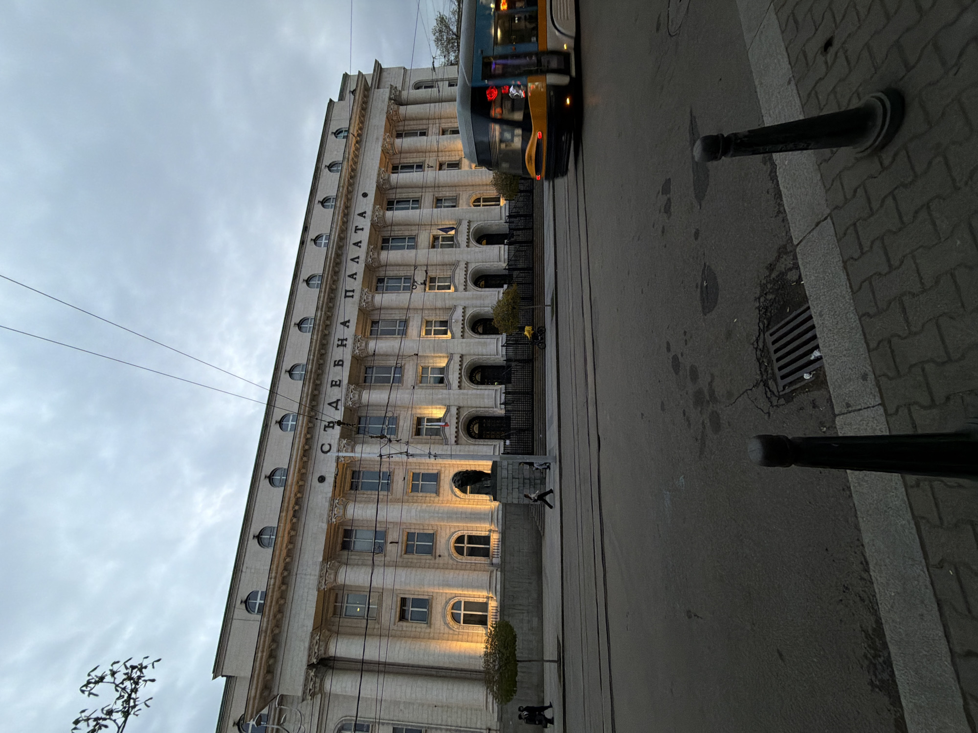 The image shows the Palace of Justice in Sofia, Bulgaria. The building is a large, imposing structure with a classical facade. A tram is partially visible on the right side of the frame. There are black bollards on the sidewalk to the right of the tram. People are seen walking in front of the building and a small tree is situated in front of the lower section. The sky is overcast.