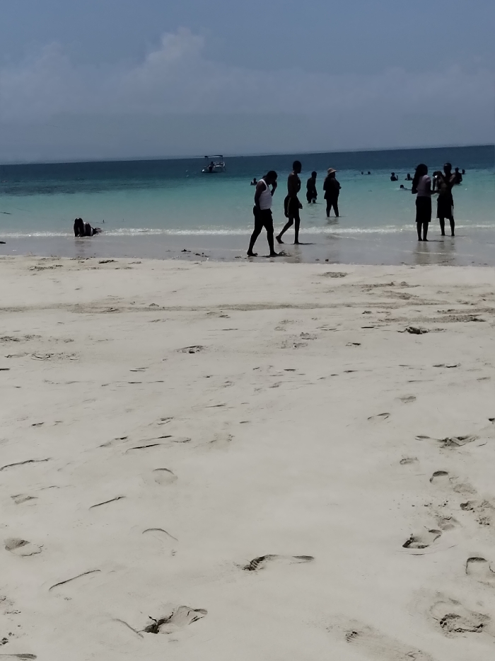 The image depicts a bright, sunny day on a white sandy beach in Wasini, Kenya. The foreground consists of light-colored sand with numerous visible footprints and minor textural variations. Moving towards the midground, the sand transitions into clear, shallow turquoise ocean water.
Several individuals are present. In the shallow water on the far left, one person is partially submerged or kneeling. Closer to the center, a male figure in a white tank top and dark shorts walks towards the right, partially obscured by another silhouetted male figure walking in the same direction. Further right, a small group of three people stands in the shallow water; two are largely silhouetted and appear to be conversing or facing each other, while a third stands slightly behind them, facing away. Additional indistinct figures are visible further out in the water, seemingly swimming or wading. A small motorboat or dinghy with occupants is positioned in the mid-distance on the water, slightly to the left. The background features open ocean meeting a clear horizon under a pale blue sky with scattered white clouds. The overall scene shows people engaged in leisure activities on a coastal environment.