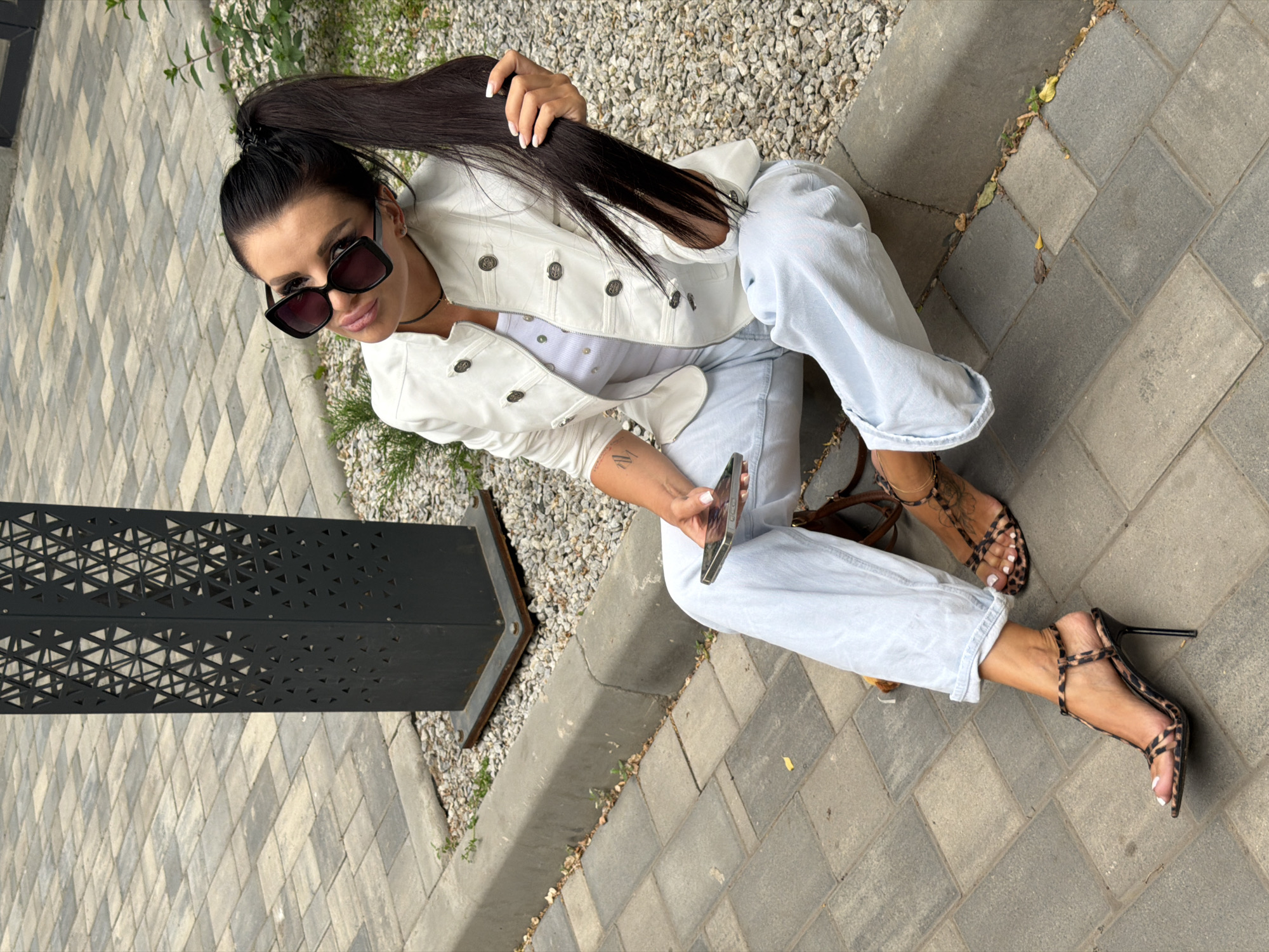 A woman is sitting on a sidewalk in front of a lamppost with a decorative geometric pattern. She is wearing a white jacket, a white top, light blue jeans, and leopard-print high-heeled sandals. She has dark hair pulled back into a ponytail, wears large sunglasses, and holds a smartphone in her hand. The ground is paved with interlocking bricks and gravel. The scene appears to be in an urban or suburban outdoor setting, possibly in Novaya Usman', Russia.