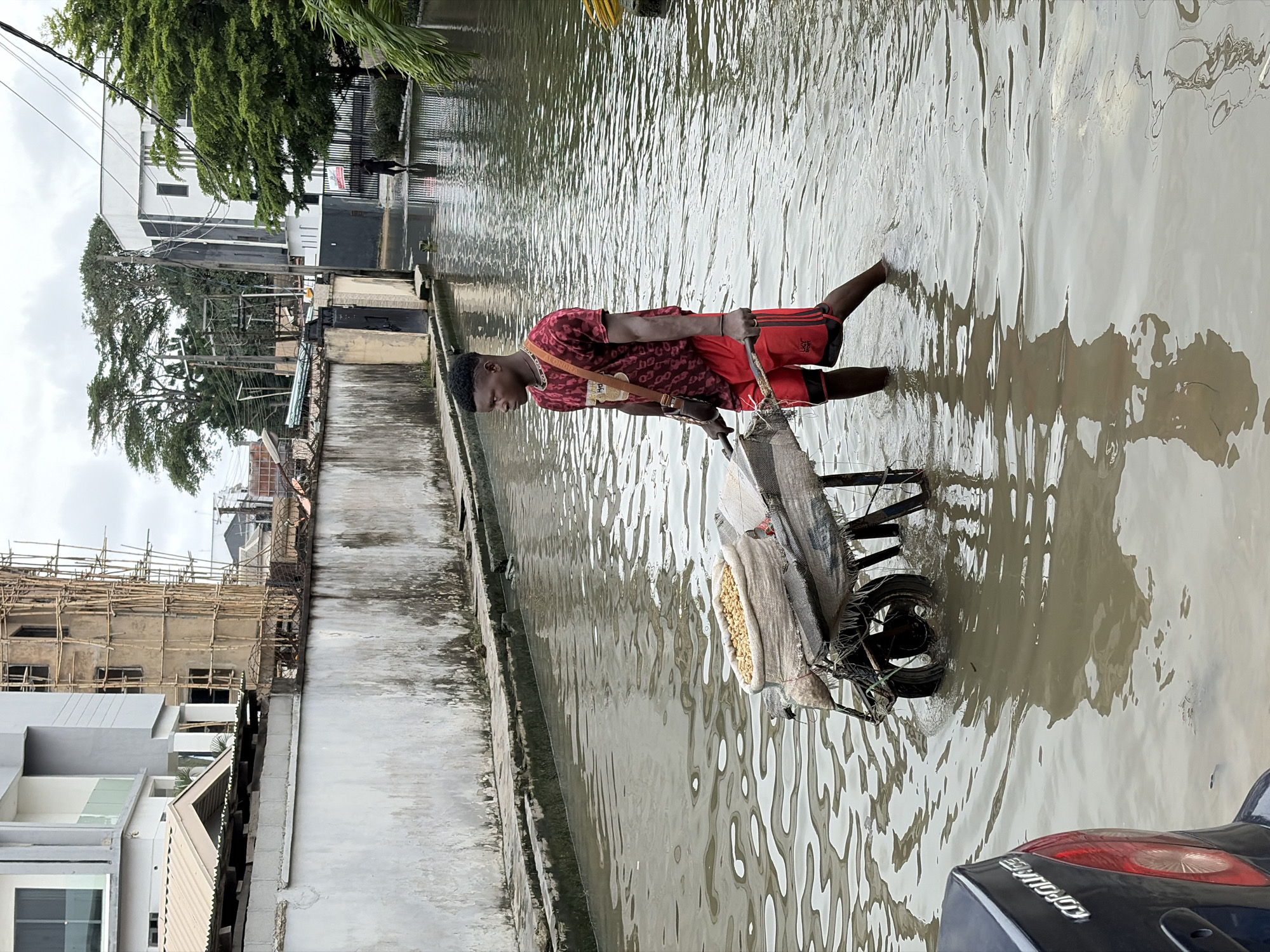 A young Black male is walking through flooded streets, pushing a wheelbarrow. He is wearing a red t-shirt with a black leopard print and red shorts with black stripes. A brown strap is slung across his chest, and a gold chain is visible around his neck. The wheelbarrow contains a large sack filled with what appears to be yellow corn kernels. The scene is set in a residential or commercial area with buildings lining the flooded street. Construction scaffolding is visible on one of the buildings, suggesting ongoing development. The water level is high enough to submerge the lower parts of the wheelbarrow's wheel and the man's legs up to his knees. The sky is overcast, indicating a cloudy day. The location is Igboefon, Nigeria.