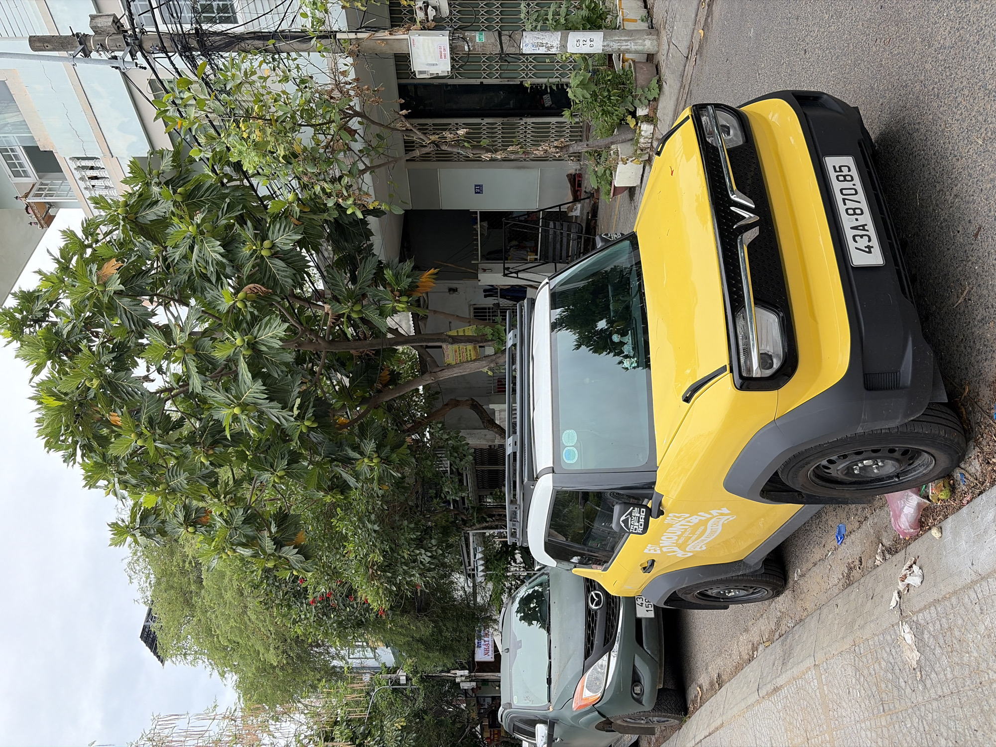 The image captures a street scene in Da Nang, Vietnam. In the foreground, a bright yellow electric car with a black roof rack and black wheel arches is parked on the right side of the road. The car has a "W" logo on its front grille and a license plate that reads "43A-870.85". A sticker on the side mirror displays "TRAVEL OFF ROAD" and "EST OLD MOUNTAIN 2023". To the left of the yellow car, a dark green SUV is parked. This SUV has a license plate that reads "43 156". Both vehicles are parked on a paved street with a concrete sidewalk on the left. The sidewalk is lined with some debris.
The background features lush green trees, some bearing fruit, which partially obscure the buildings. A light blue, multi-story building with balconies is visible on the right side of the image, behind the yellow car. Electrical poles and numerous wires are also prominent. The sky is overcast. The scene appears to be in a residential or commercial area of Da Nang, likely a street with businesses and homes.