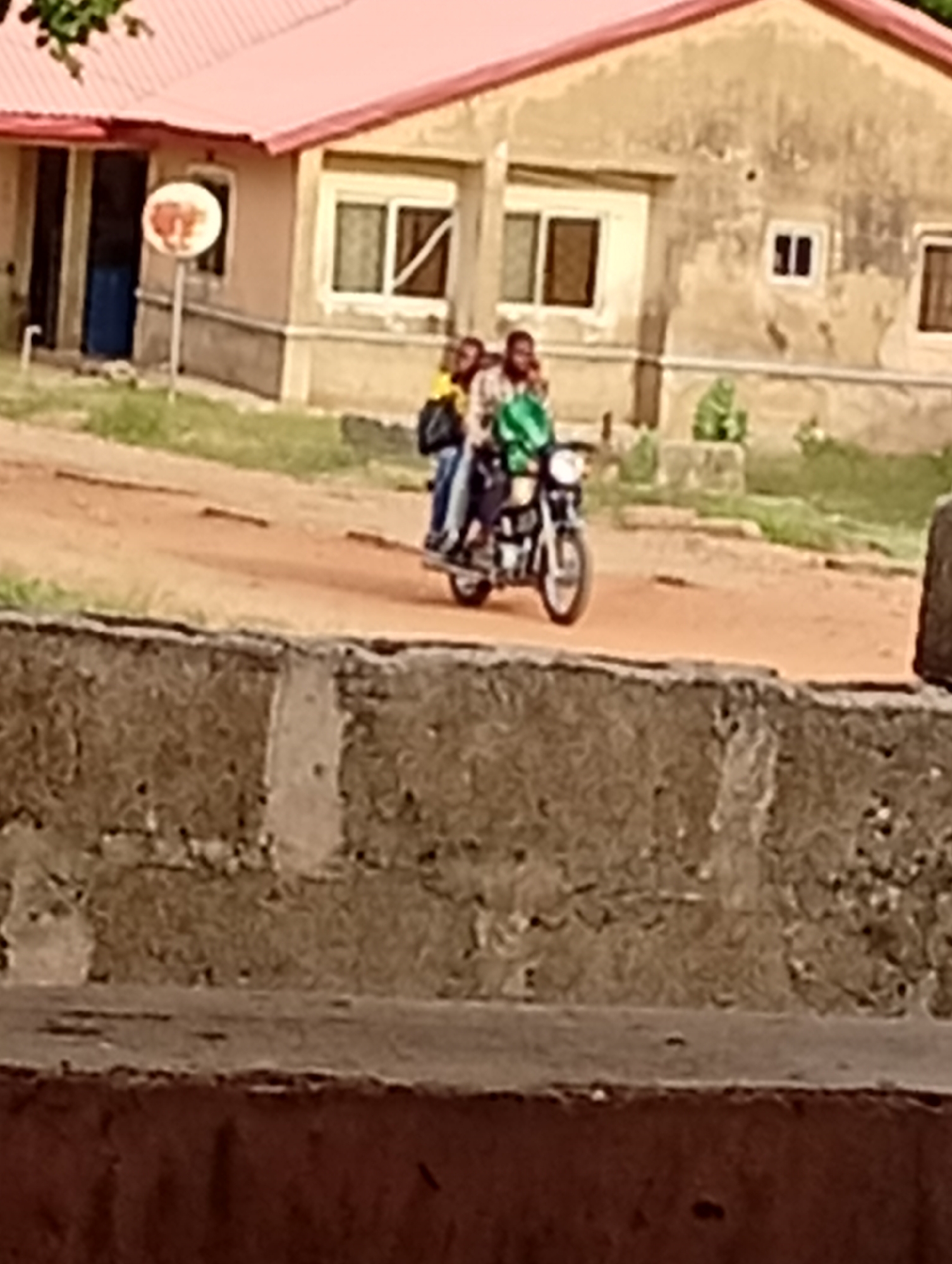 The image depicts an outdoor scene featuring three people on a motorcycle on an unpaved road. The motorcycle is positioned on a reddish-brown dirt or gravel path. The driver wears a green long-sleeved shirt, and two passengers are seated behind, one wearing a yellow top and the other partially obscured.
In the background, a single-story building with light-colored (beige or yellowish) walls and a reddish-orange corrugated metal roof stands. The building appears weathered, with several windows, some of which may have security bars or shutters, and a prominent blue door on its left side. Adjacent to the blue door, a white circular sign with an indistinct orange or reddish design/logo is mounted on a pole.
The foreground shows a textured concrete block wall or fence, also exhibiting signs of weathering. Some green foliage is visible in the upper left corner and sparse grass lines the roadside. The lighting is soft and diffused, suggesting daytime, possibly under an overcast sky or during the late afternoon. The setting appears to be a rural or semi-urban environment. No discernable text is visible.