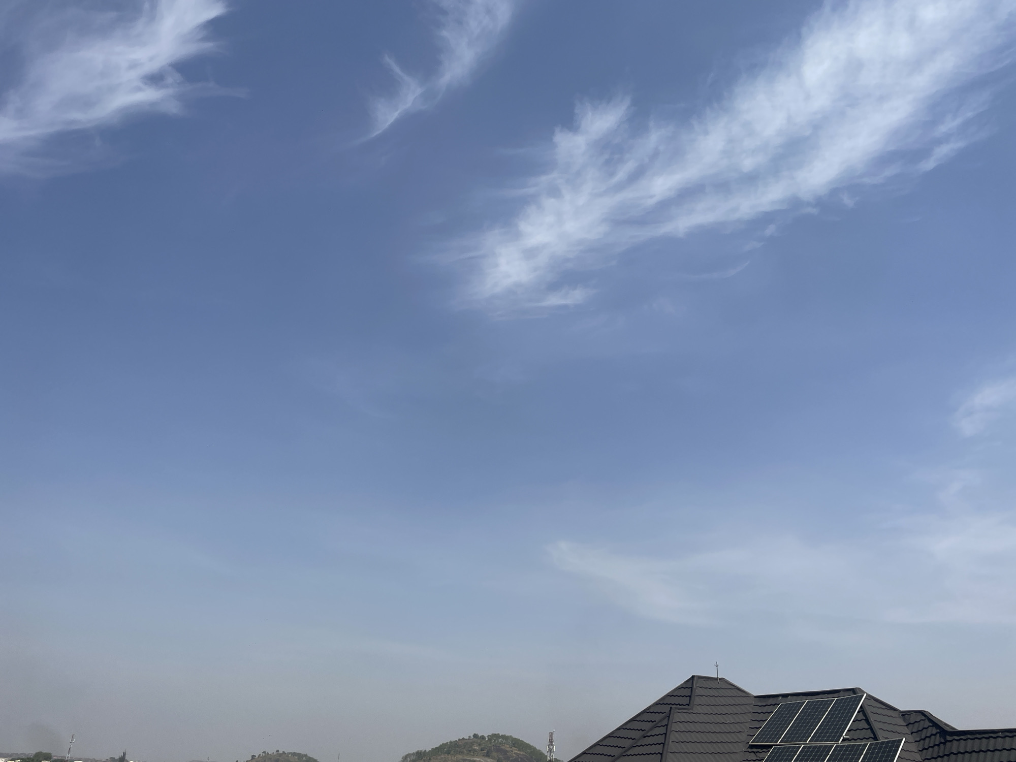 The image shows a view of the sky above Abuja, Nigeria. In the upper part of the frame, wispy cirrus clouds are scattered across a pale blue sky. The lower portion of the image includes a section of a dark gray tiled roof with solar panels installed. The horizon shows a hazy atmospheric condition with suggestions of low hills or small mountains in the distance. At the left, there's a vague suggestion of buildings or structures partially obscured by the atmospheric haze.