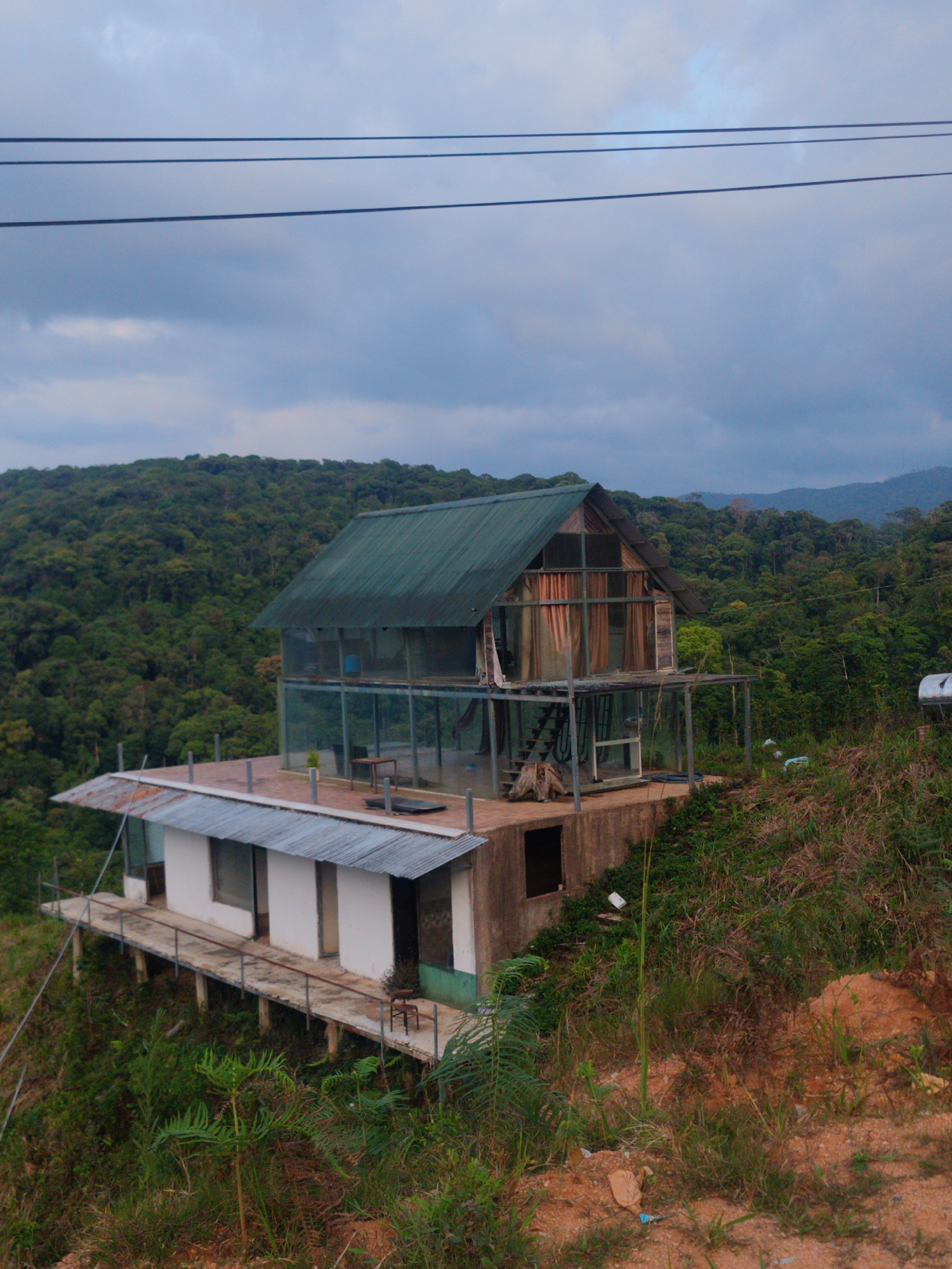 A two-story structure with a mix of construction styles sits on a hillside in Dam Rong 4, Vietnam. The lower level features white walls with grey trim and a flat grey metal roof extending out, supported by concrete columns. A small chair sits on the extended platform. The upper level has a pitched green metal roof. The walls appear to be made of a frame with transparent panels. A set of stairs can be seen through the panels. The building is surrounded by dense green forest. Power lines run across the top of the image. The ground is covered in grass and dirt.