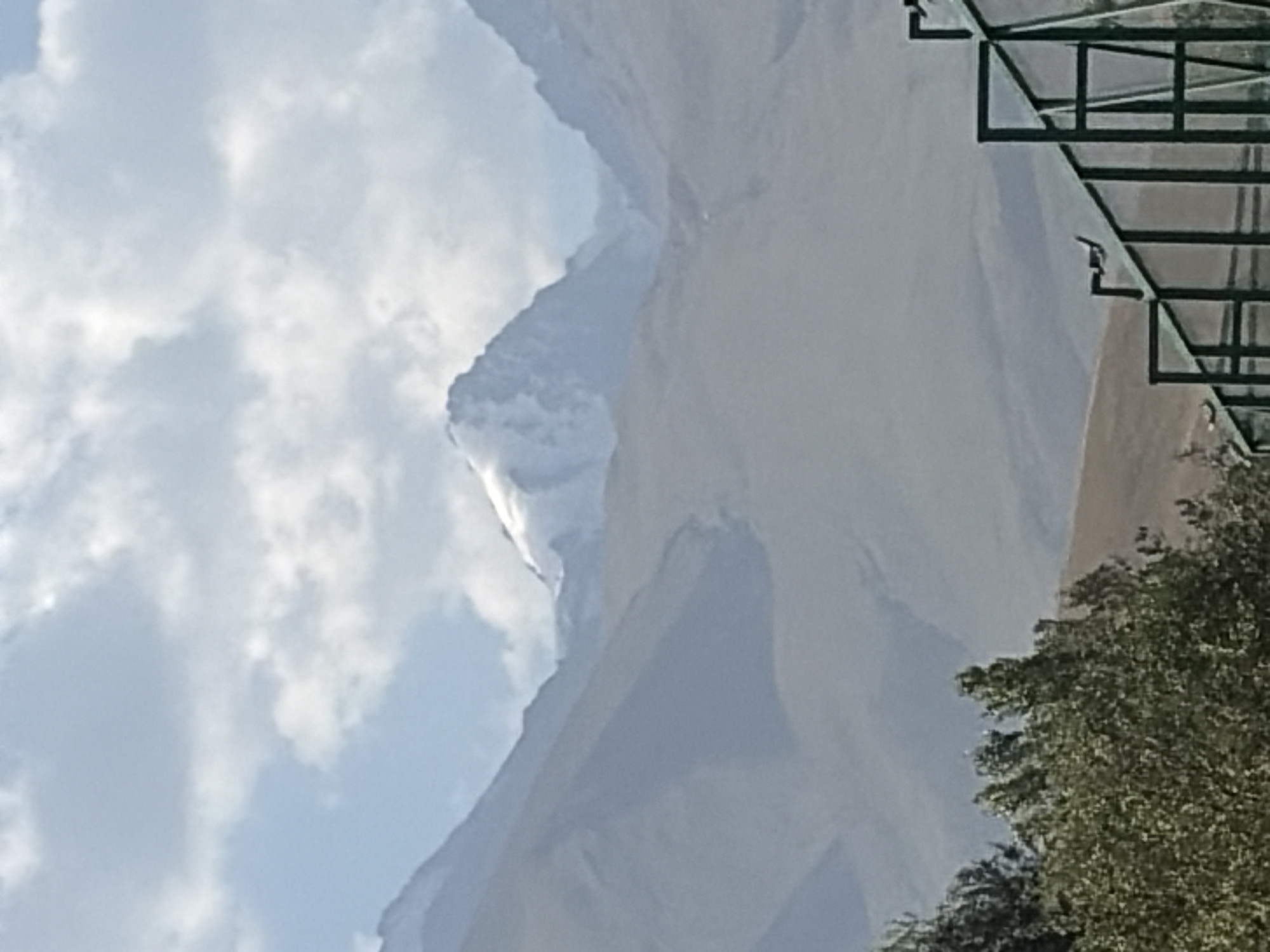 This is a photograph of a mountainous landscape with a clear sky and scattered clouds. In the foreground, there is a green metal fence with glass panels. Behind the fence, there are trees with green foliage. The middle ground features rolling hills and mountains. In the background, a prominent snow-capped mountain stands tall, with other peaks visible on either side. The lighting suggests it is daytime. The location is described as Unknown, Unknown.