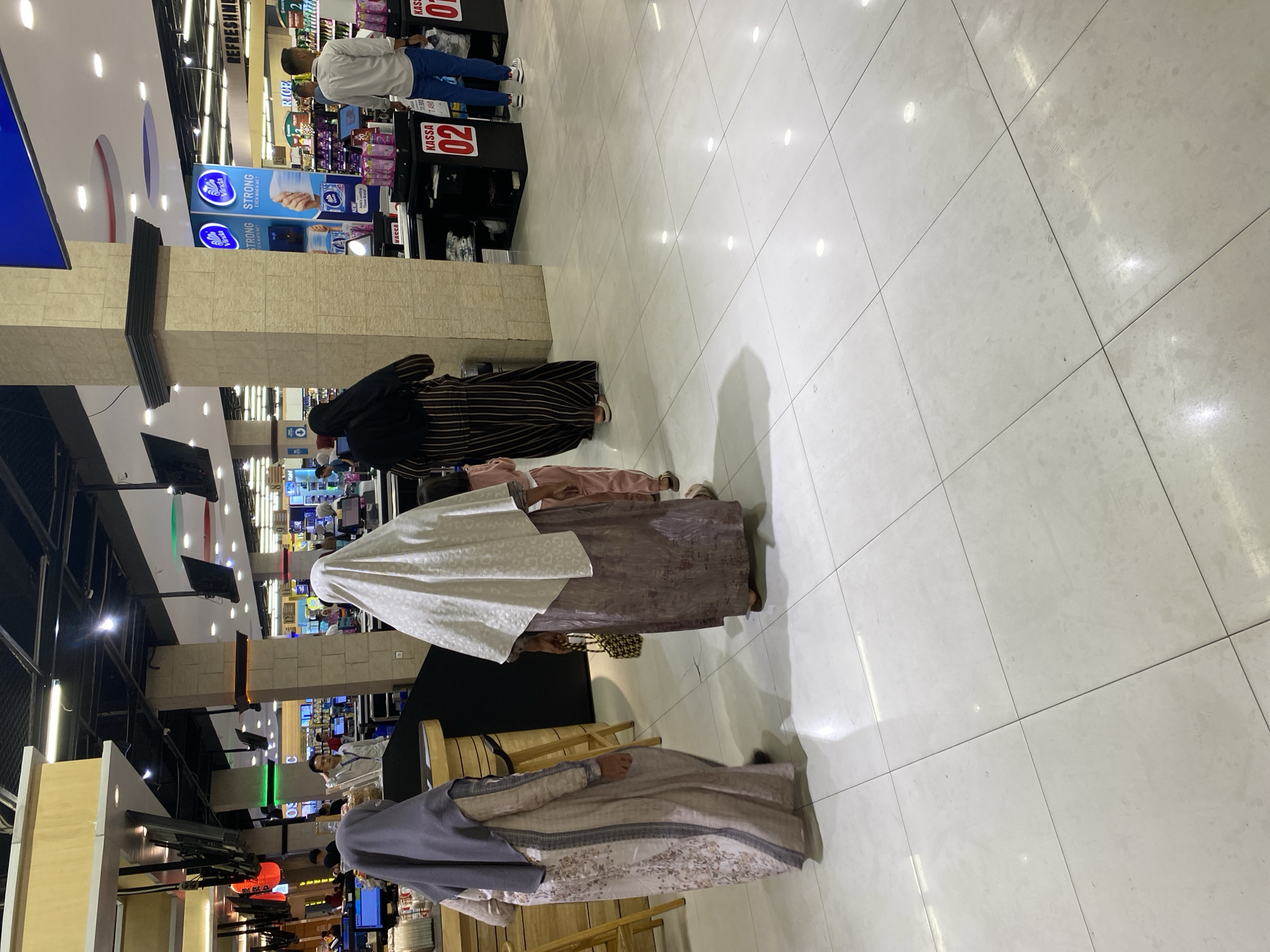 In the interior of a brightly lit retail store, likely a department store or supermarket, in Banda Aceh, Indonesia, several individuals are observed near checkout counters. Three women, two of whom are wearing hijabs, are the central focus. One woman, on the left, wears a light-colored, patterned dress and a dark grey hijab. She is positioned behind a wooden counter. To her right, another woman, also in a hijab, wears a dark striped abaya. She is holding a small, woven bag and is facing away from the camera. In front of her, a young child in pink trousers and a light pink top is partially visible. A third woman, on the far right, wears a white hijab and a purple-toned dress, also facing away. She appears to be walking past a checkout counter labeled "KASSA 02". Further to the right, a man in a light blue shirt and blue trousers stands near a checkout counter labeled "KASSA 01".
The environment features a polished tiled floor that reflects the overhead lighting. The background reveals displays of merchandise, promotional signage, and ceiling structures with lighting and ventilation systems. The checkout counters are equipped with electronic systems and payment terminals. The overall scene depicts a typical retail environment during operating hours, with customers engaged in shopping and checkout processes. The presence of specific signage like "KASSA 01" and "KASSA 02" indicates this is likely a cashier station in a retail setting.