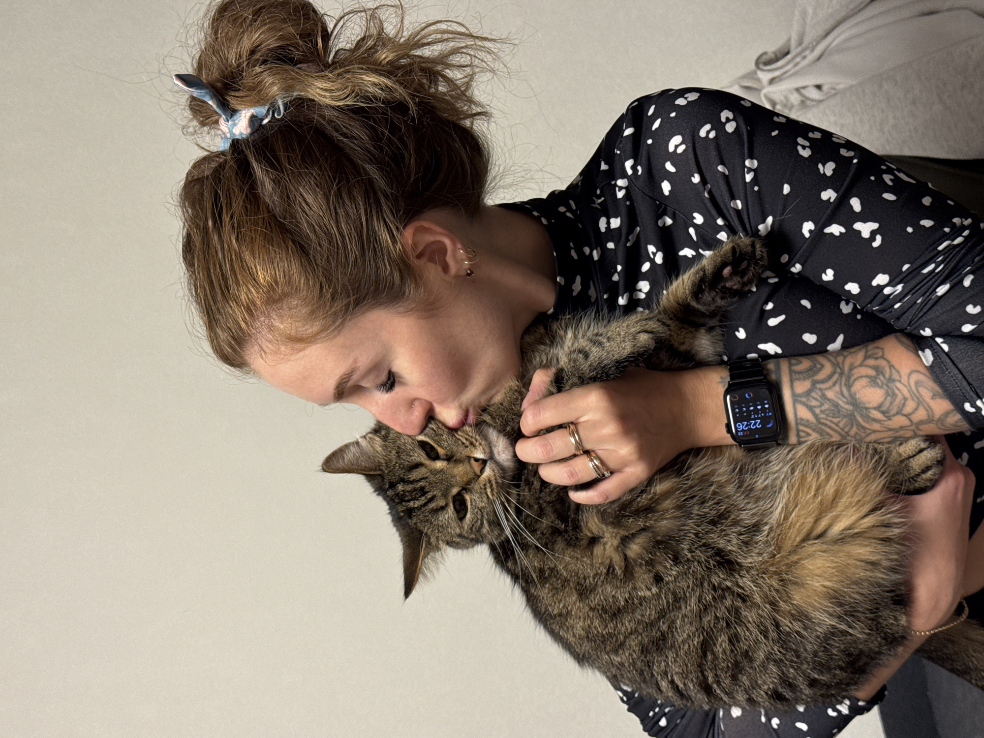 An affectionate moment between a woman and her tabby cat is captured in an indoor setting. The woman, with her light brown hair styled in a messy bun secured by a patterned hair claw, is seen holding the cat close and gently kissing its forehead. She is wearing a black long-sleeved shirt adorned with small white, irregular dot patterns.
Notable details of the woman include several gold rings on her right hand, a gold hoop earring with a small red drop, and a small stud piercing above her lip. On her left forearm, a detailed black and grey tattoo is visible. Her left wrist displays an Apple Watch showing the time as "22:26," a temperature of "0.2°C," and a moon icon, indicating late evening or night. The text "ЧТ 23" is also visible, likely referring to "Thursday 23" in Cyrillic, suggesting a date and cold weather consistent with regions like Chaikovsky, Russia.
The cat is a fluffy, brown and black striped tabby with prominent white whiskers. It appears calm and comfortable in the woman's embrace. The background consists primarily of a plain, light-colored wall, with a folded light grey textile or blanket visible in the upper right corner, indicating a cozy, domestic environment. The image conveys a strong bond and tenderness between the pet and its owner.