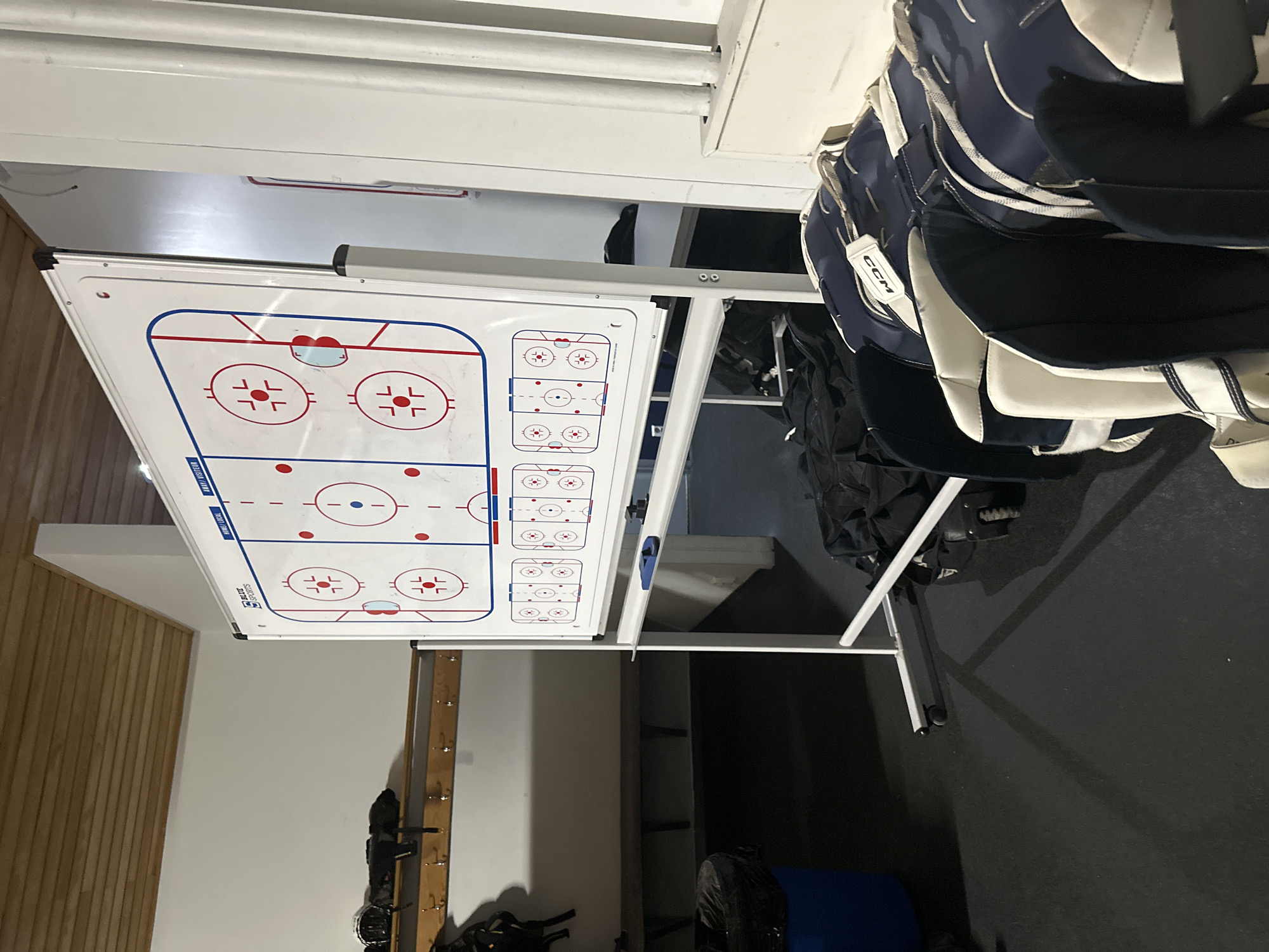 The image shows a hockey whiteboard on a stand in what appears to be a locker room or training facility in Unknown, Unknown. The whiteboard displays a diagram of a hockey rink with lines and face-off circles. A rack of hockey equipment is visible to the right of the whiteboard. The environment appears to be indoor and functional.
