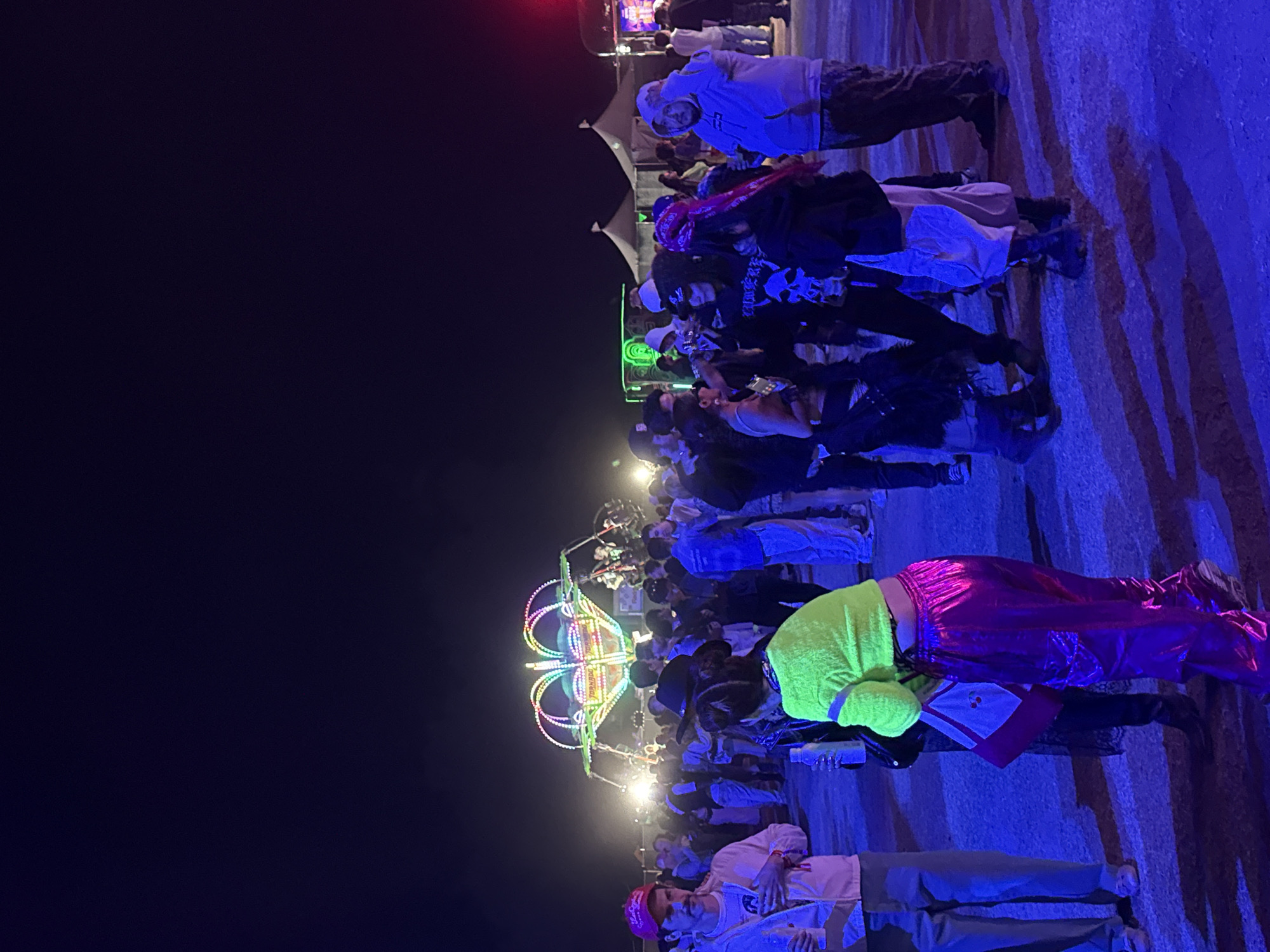 This image depicts a nighttime scene in Thermal, California, United States of America. A crowd of people is gathered near a brightly lit carnival ride. The environment is illuminated by various colored lights, including blue lighting across the ground, and a green light at the top left of the crowd. A person in a neon green top and shiny pink pants is visible in the foreground. Tents can be seen in the background, beyond the crowd. People appear to be interacting and enjoying the carnival.