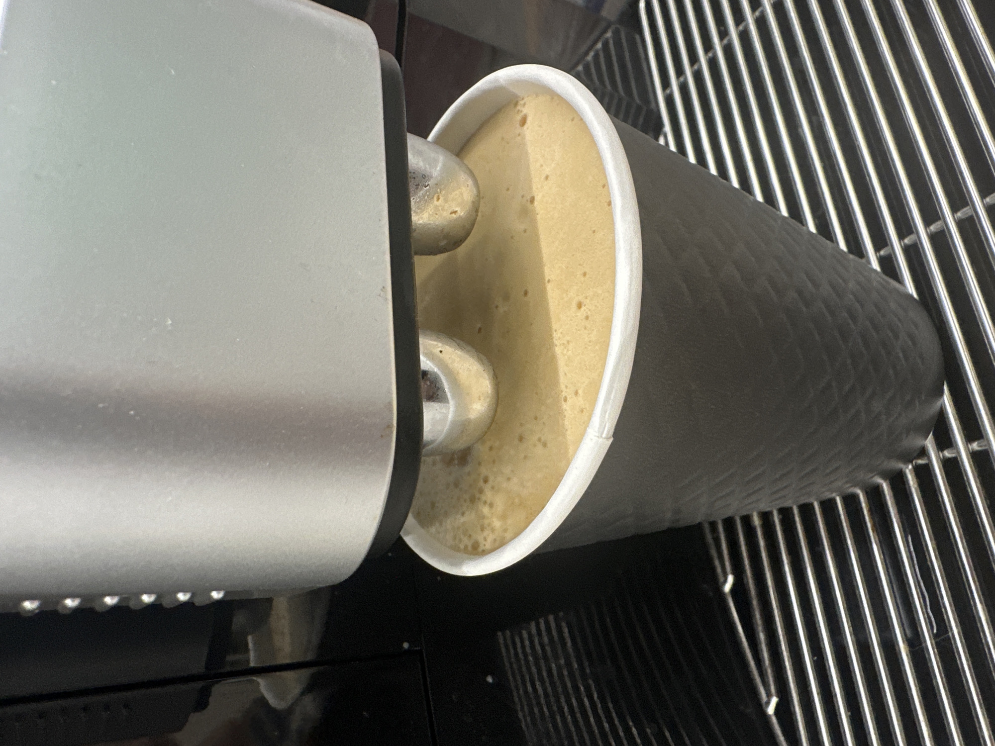 A close-up, high-angle shot depicts a light brown beverage with a foamy surface being dispensed into a black paper cup with a white rim from a silver machine. The cup is resting on a metal grate. The beverage is a light coffee drink. The setting may be in a coffee shop. There is no evidence to indicate the location in Бараўлянскі сельскі Савет, Belarus.