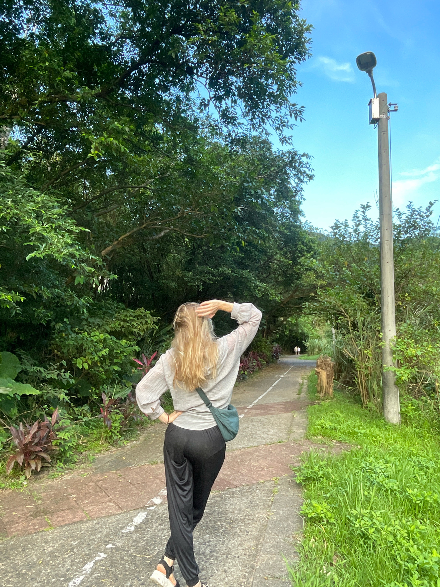 A blonde woman wearing a light-colored long-sleeved shirt, black pants, and sandals is captured from behind, walking away from the camera down a paved path in a park. She has one hand on her hip and the other is raised to her head, adjusting her hair. A green crossbody bag is slung over her left shoulder. The path is lined with lush green trees and vegetation, with a street light pole on the right side of the frame. The sky above is blue with a few wispy clouds. In the distance, two small figures can be seen walking further down the path. The setting appears to be a park or trail, possibly in New Taipei City, Taiwan, given the presence of dense foliage and the overall atmosphere of a public outdoor space.