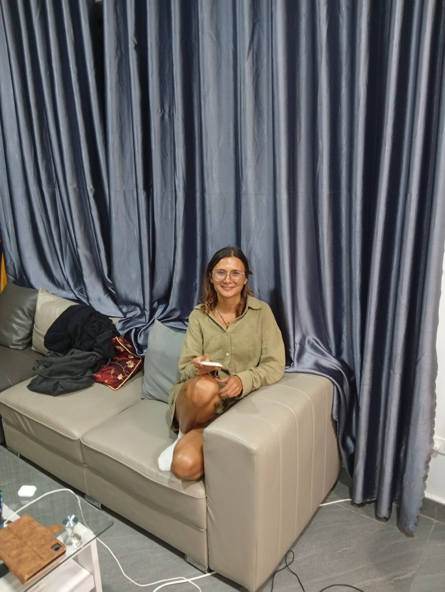 The image shows a woman sitting on a light beige couch in front of a large, floor-to-ceiling gray curtain. The woman is wearing glasses and a button-down olive green shirt. She's holding a white smartphone. On the couch next to her are a gray pillow, a black garment, and a patterned cushion. A glass-topped coffee table is visible in the lower left corner, with a brown tablet and charging cables on it. The flooring is gray and tile-like. A power outlet is visible on the wall to the right of the couch. The image was taken in Da Lat, Vietnam.