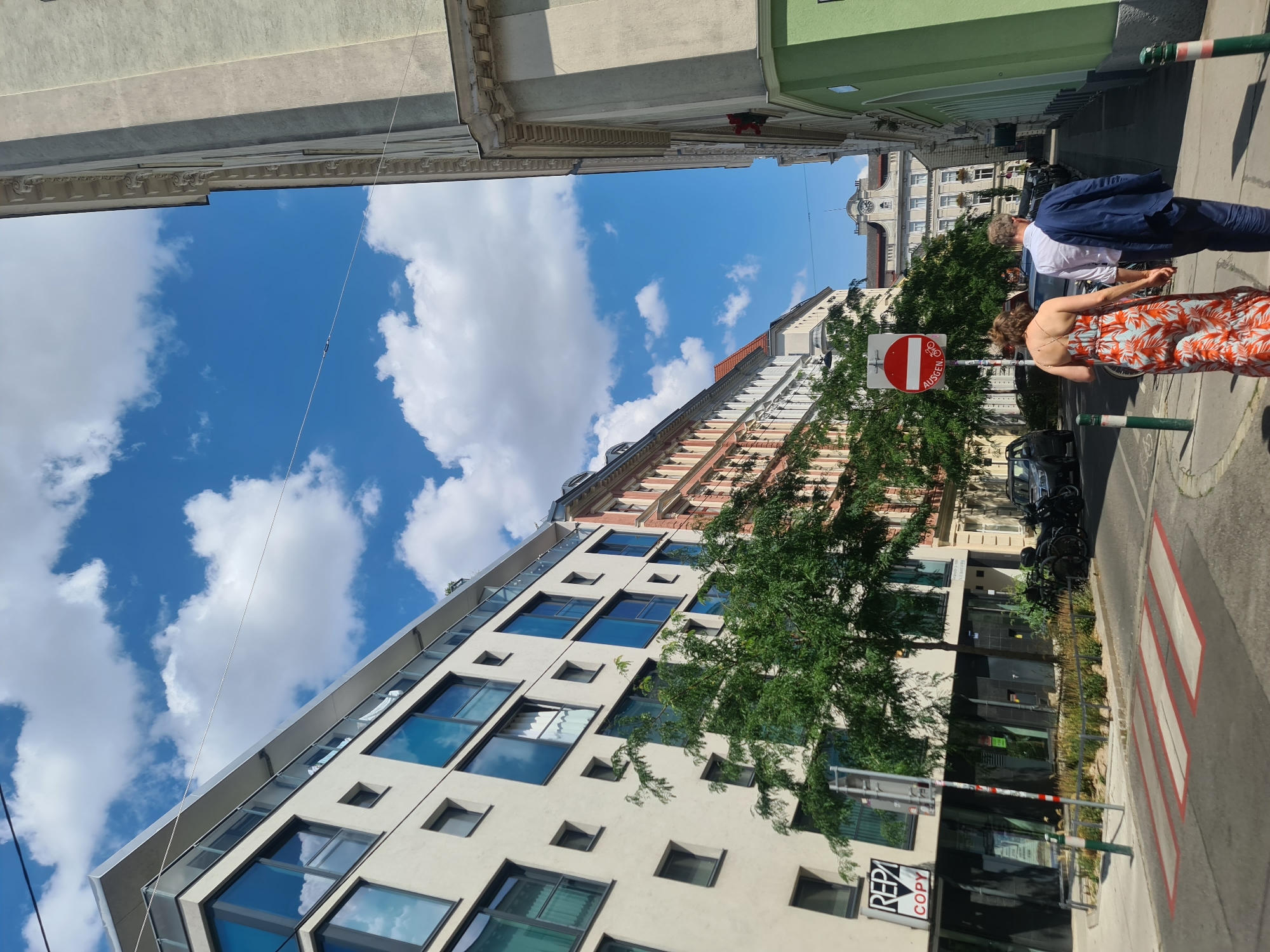 The image depicts a street scene in Vienna, Austria, on a sunny day with scattered clouds. In the foreground, a pedestrian crossing with red and white markings is visible. To the left, a modern white building with large blue-tinted windows and balconies stands prominently. A sign with "REPA COPY" is attached to its facade. Beside this building, a tree with lush green foliage grows. Further down the street, a row of older buildings with colorful facades, predominantly orange and white, are seen. On the right side of the street, a tall, light-colored building with ornate architectural details lines the path. Further into the distance, more buildings and a clock tower are visible. Two people are walking away from the camera down the street. The person on the left is wearing a long, brightly colored floral dress, and the person on the right is wearing a white shirt and dark trousers, with a dark blue jacket draped over their shoulders. Several parked cars and motorcycles are also present along the street. A "No Entry" sign with German text ("AUSGEN") is mounted on a pole, also featuring a bicycle symbol. The overall impression is of a typical urban street in Vienna during daytime.