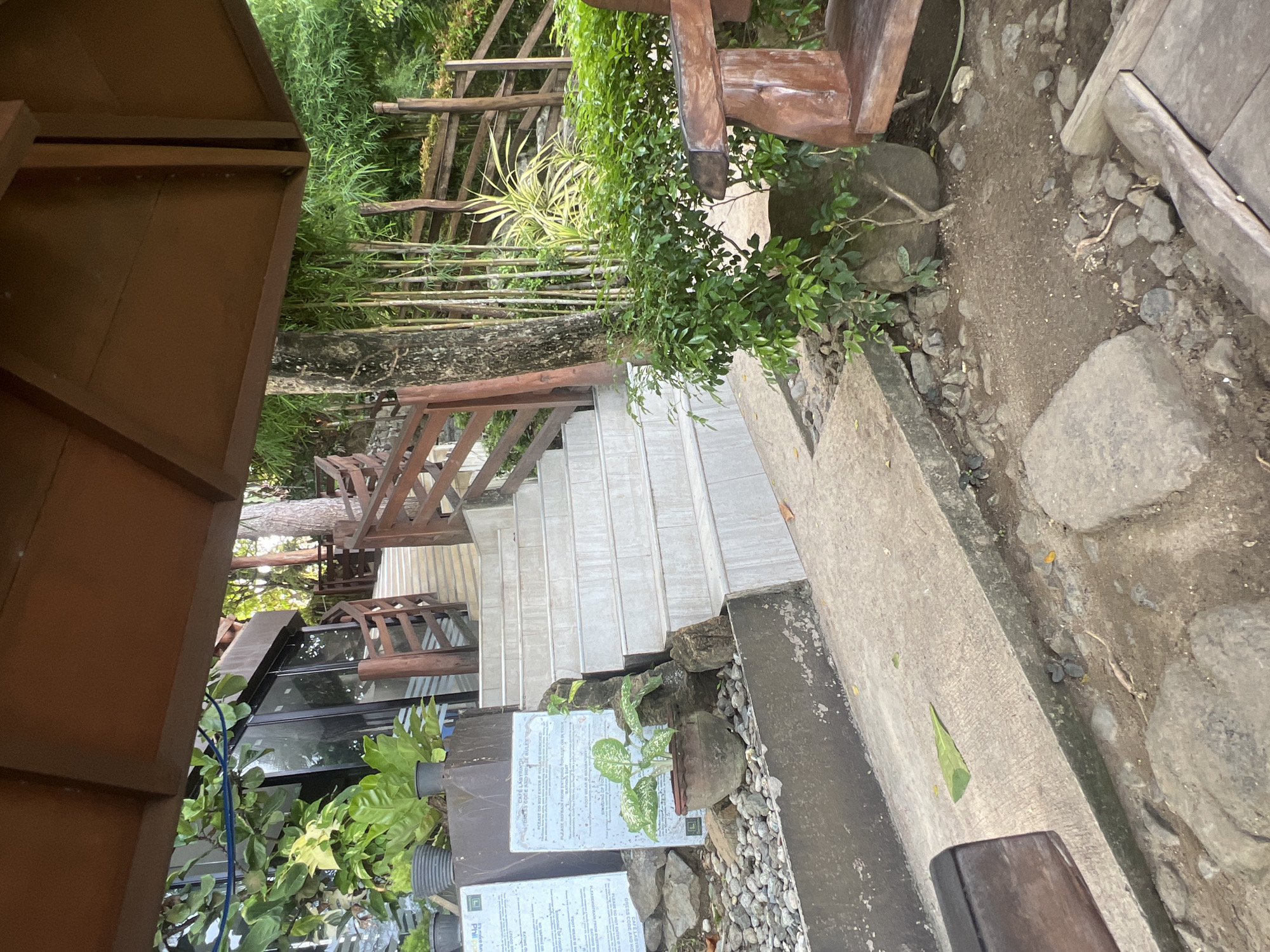 The image depicts a scene in Banaybanay, Philippines, focused on a set of concrete stairs leading upwards. The steps are white or light gray, and framed by a textured concrete border on the right. A wooden railing is visible along the staircase, partially obscured by foliage. A variety of plants, including some in pots, are positioned near the base of the stairs. Signage is posted. A wooden chair is located near the stairs. The ground around the stairs consists of a mixture of dirt, pebbles, and rocks. A wooden structure is seen on the left of the frame. The overall environment suggests an outdoor setting, possibly a garden or resort area. The scene is photographed from a slightly tilted perspective.