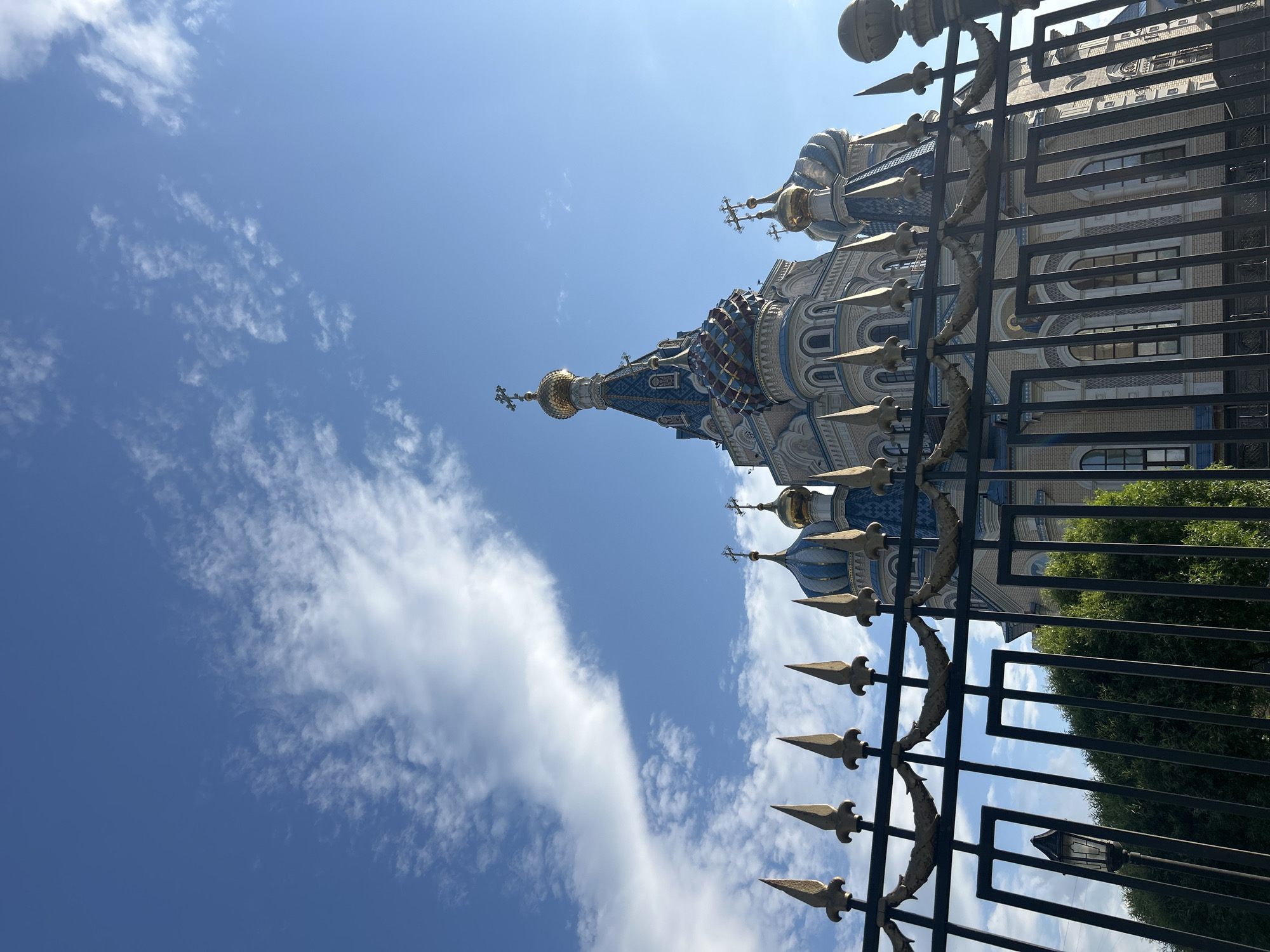 A low-angle shot of the exterior of the Church of the Assumption in Izhevsk, Russia, partially obscured by a metal fence. The church features traditional Russian Orthodox architecture, including a tall, ornate central tower and several smaller domes, all adorned with gold crosses. The building is painted in a combination of white, blue, and gold colors. A decorative metal fence runs in front of the church, casting shadows on the building. The sky is blue with scattered white clouds. Green foliage is visible behind the fence in the lower right of the frame.