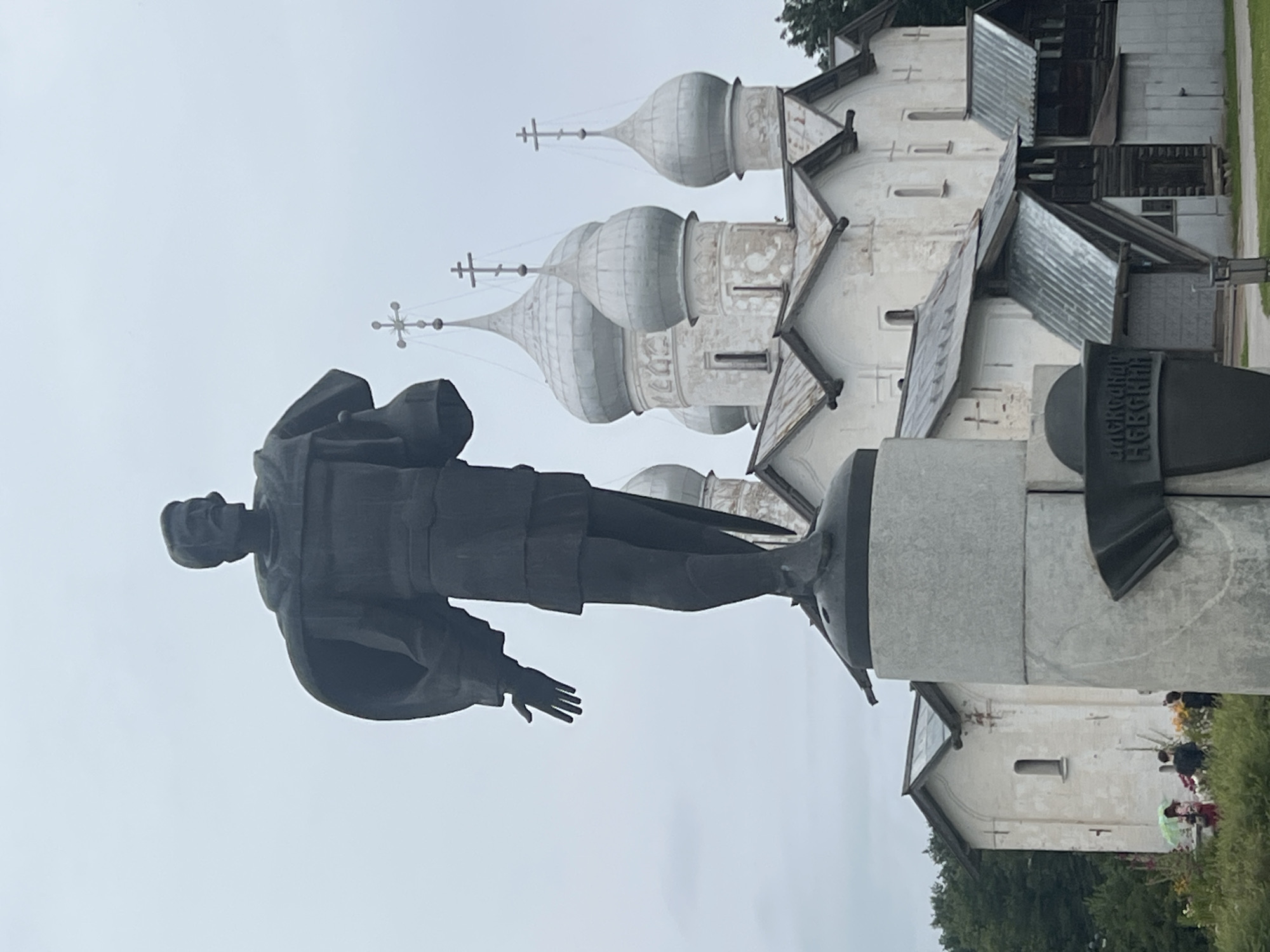 The photograph captures a statue of Prince Yaroslav the Wise in Veliky Novgorod, Russia, standing prominently in front of the Saint Sophia Cathedral. The statue, rendered in dark bronze, depicts the prince in a cloak and tunic, with his left arm extended forward. He sports a mustache and a distinctive hairstyle. Behind the statue, the white stone walls and silver onion domes of the cathedral are visible. The sky is overcast, casting a diffused light on the scene. In the foreground, at the base of the statue, a bronze inscription plate is affixed to a stone pedestal. To the left and further back, a small group of people are visible amidst greenery and flowers. The overall impression is one of historical significance and architectural grandeur.