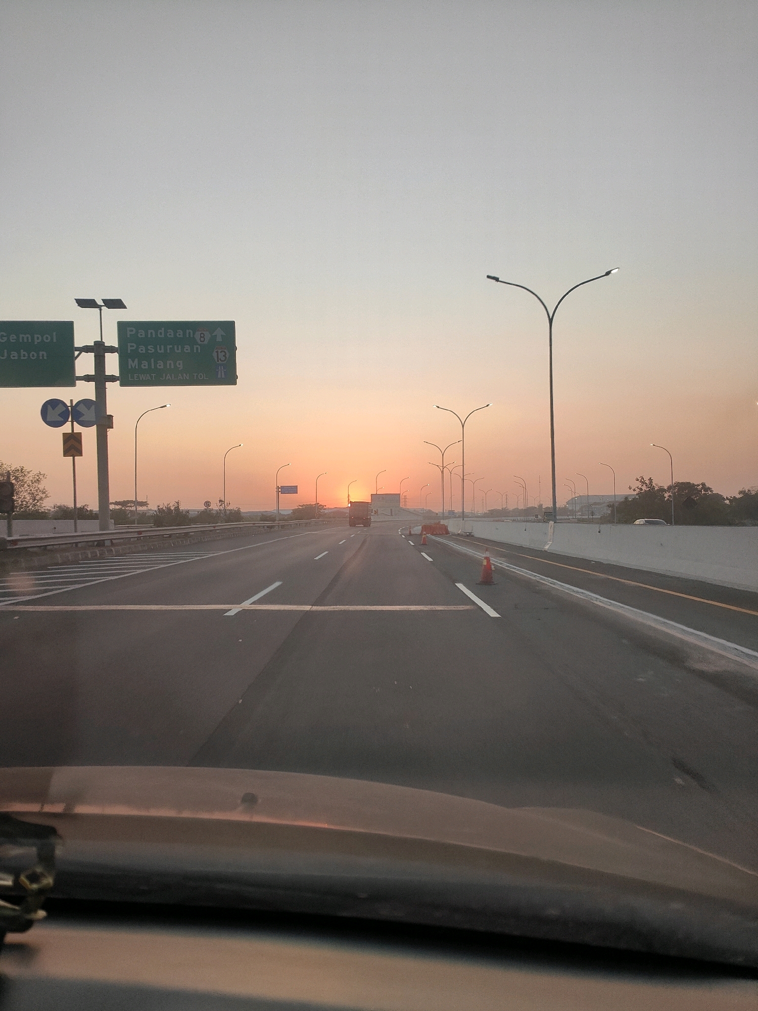 The image shows a highway scene at sunset in Gempol, Indonesia. The sky is a gradient of orange and gray, with the sun low on the horizon. A highway stretches into the distance, with multiple lanes and concrete barriers. Road signs indicate directions to Pandaan, Pasuruan, and Malang, and mention "Lewat Jalan Tol" (via toll road). Several streetlights are visible along the sides of the highway. In the foreground, the hood of a vehicle is visible, suggesting the photo was taken from a car. A truck is seen on the highway in the distance. The environment appears to be a relatively clear day with a gentle haze.