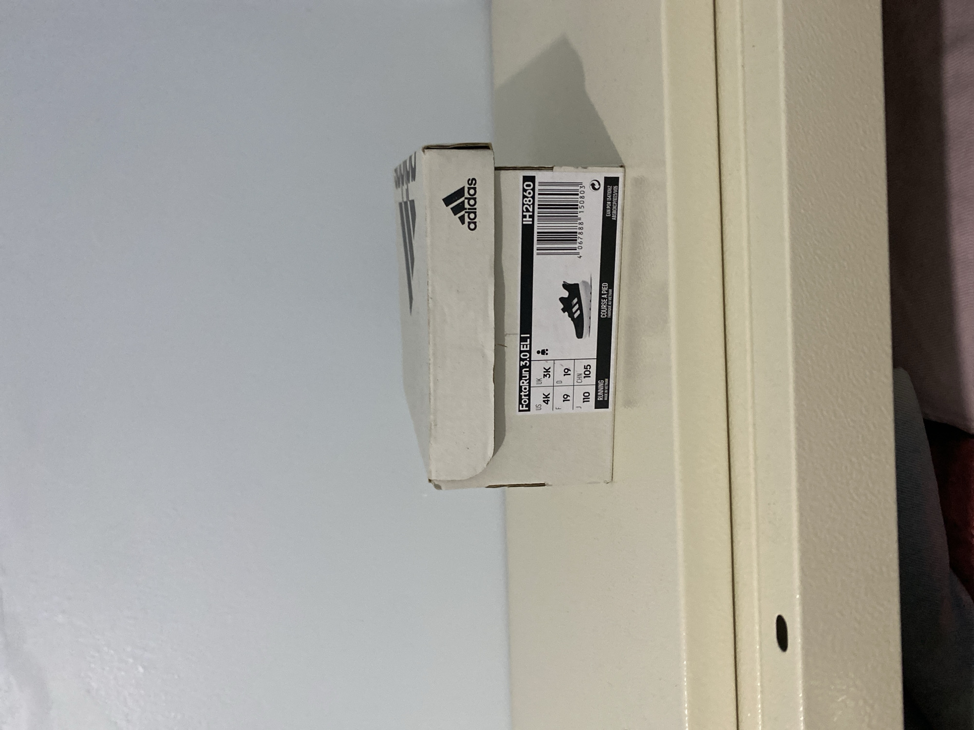 The image shows a close-up of an Adidas shoe box. The box is white with black text and the Adidas logo. The text on the box indicates that it contains "FortaRun 3.0 EL I" shoes, with sizing information in US, UK, F, D, J, and CHN. It also shows a barcode and the product code IH2860. A small silhouette of a black and white Adidas sneaker is also visible on the box. The box is sitting on a light-colored surface, possibly a shelf or dresser. The background is a plain, light-colored wall. There is no indication of the location being Dukuh, Indonesia.