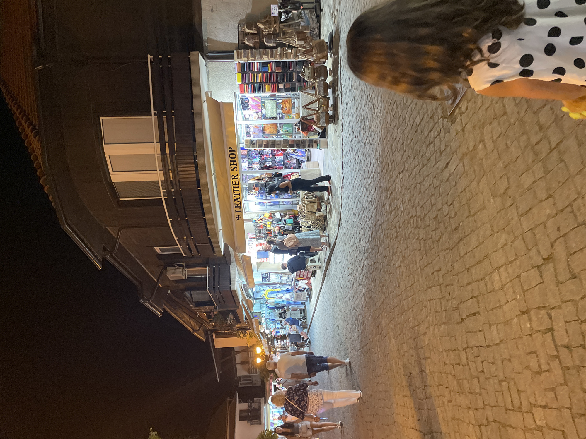 The image captures a night scene in the city of Nesebar, Bulgaria, specifically on a cobblestone street lined with shops. In the foreground, the back of a young girl with long, dark, wavy hair is visible. She is wearing a white dress with black polka dots. To her left, the cobblestone pavement is illuminated by artificial light.
The street is bustling with activity. Several people are walking along the street, while others are browsing the merchandise displayed outside the shops. On the right side of the street, a "LEATHER SHOP" is prominently advertised with a yellow awning. The shop's interior is visible, showcasing a variety of leather goods, including handbags, wallets, and belts, displayed on shelves and racks. Outside the shop, more bags are hung for display.
Further down the street, other shops are visible, selling clothing and other items. The architecture of the buildings suggests a historic European town. The overall atmosphere is lively, with people enjoying a night out in a commercial area. The lighting is dim, creating a warm, inviting ambiance.