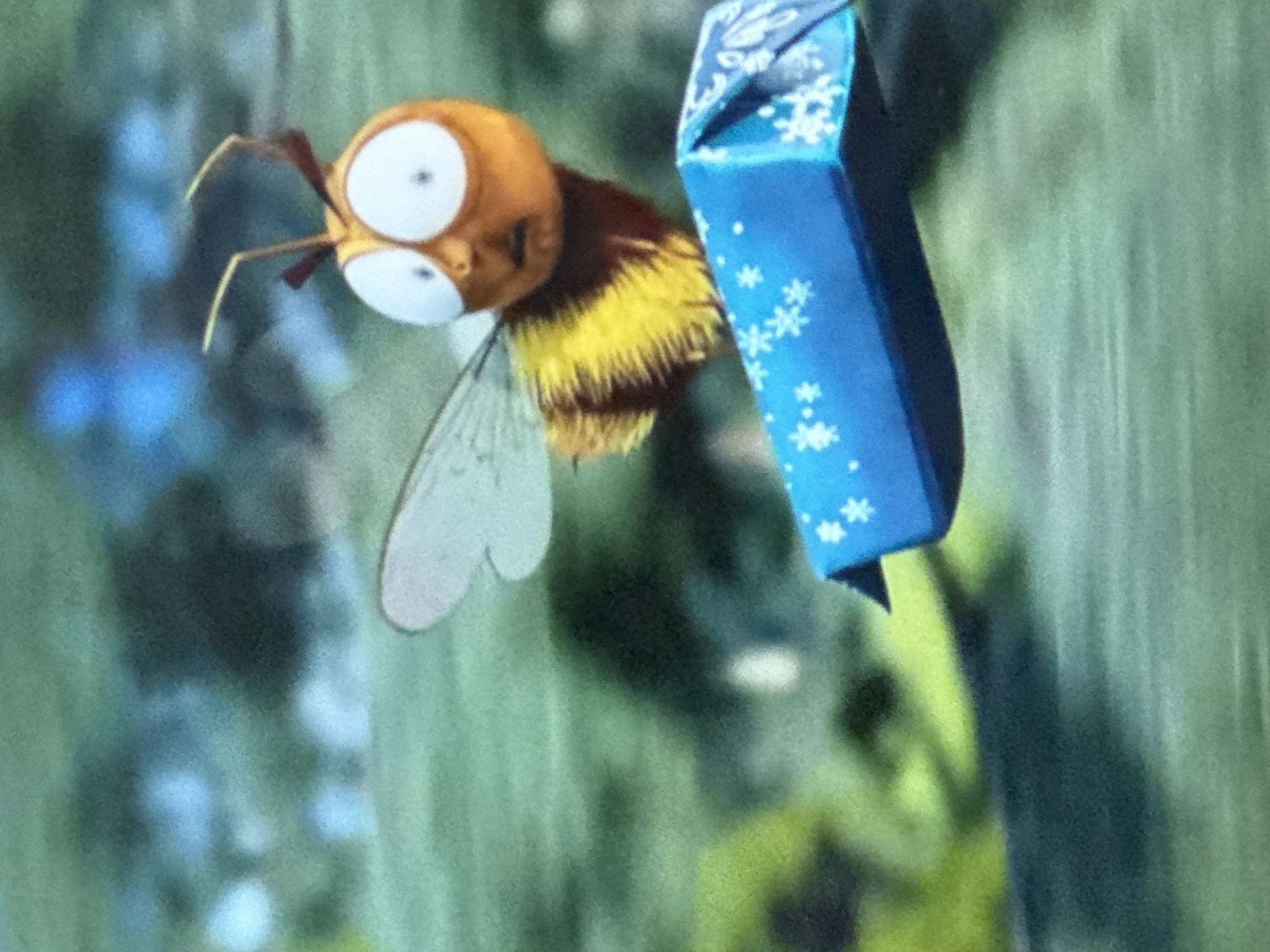 A cartoonish bee with large white eyes flies toward the left while carrying a blue, rectangular package decorated with white snowflakes. The background is blurred and appears to be green foliage.