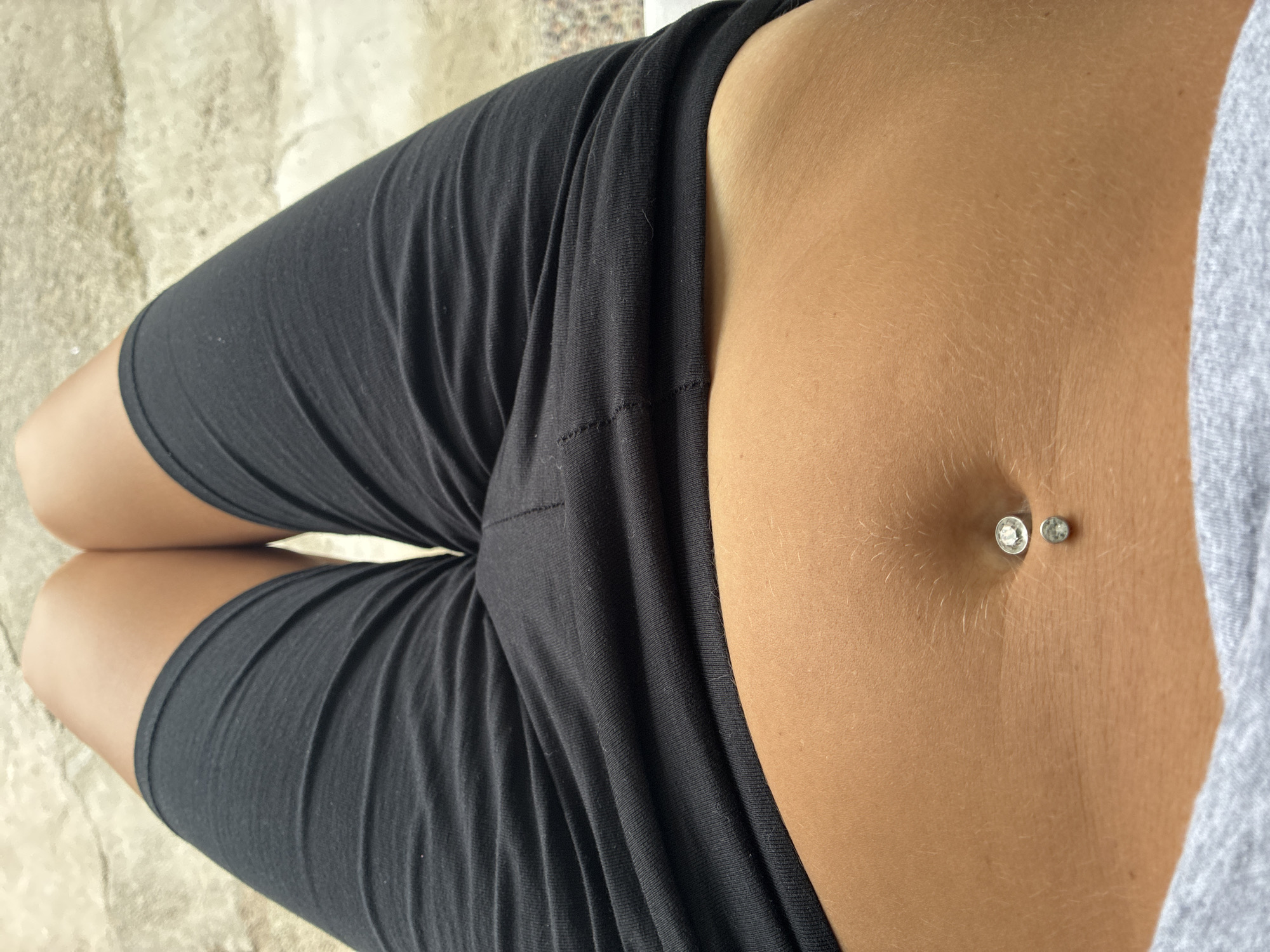 The image shows a close-up view of a person's midsection, from just above the navel to the upper thighs. The person is wearing black shorts and a grey garment, possibly a t-shirt, around the waist. The navel piercing consists of two small, sparkling jewels, one slightly larger than the other. The skin appears tanned. The background is a textured, light-colored surface, possibly concrete or stone. There is no indication of the city of Surami, Georgia, within the image.
