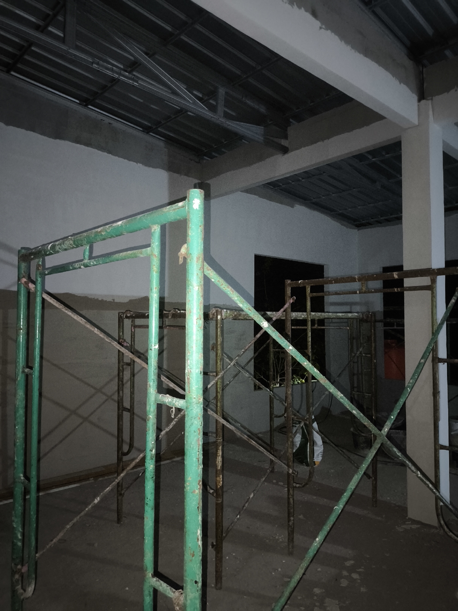 The image shows an interior construction site with scaffolding. In the foreground, green metal scaffolding is prominently displayed, showing signs of wear and tear. In the background, other scaffolding structures are visible, along with concrete walls and a metal ceiling structure. The lighting is dim, suggesting it might be evening or the interior is not fully illuminated. There are no people visible in the image. The scene depicts an unfinished building or renovation project.